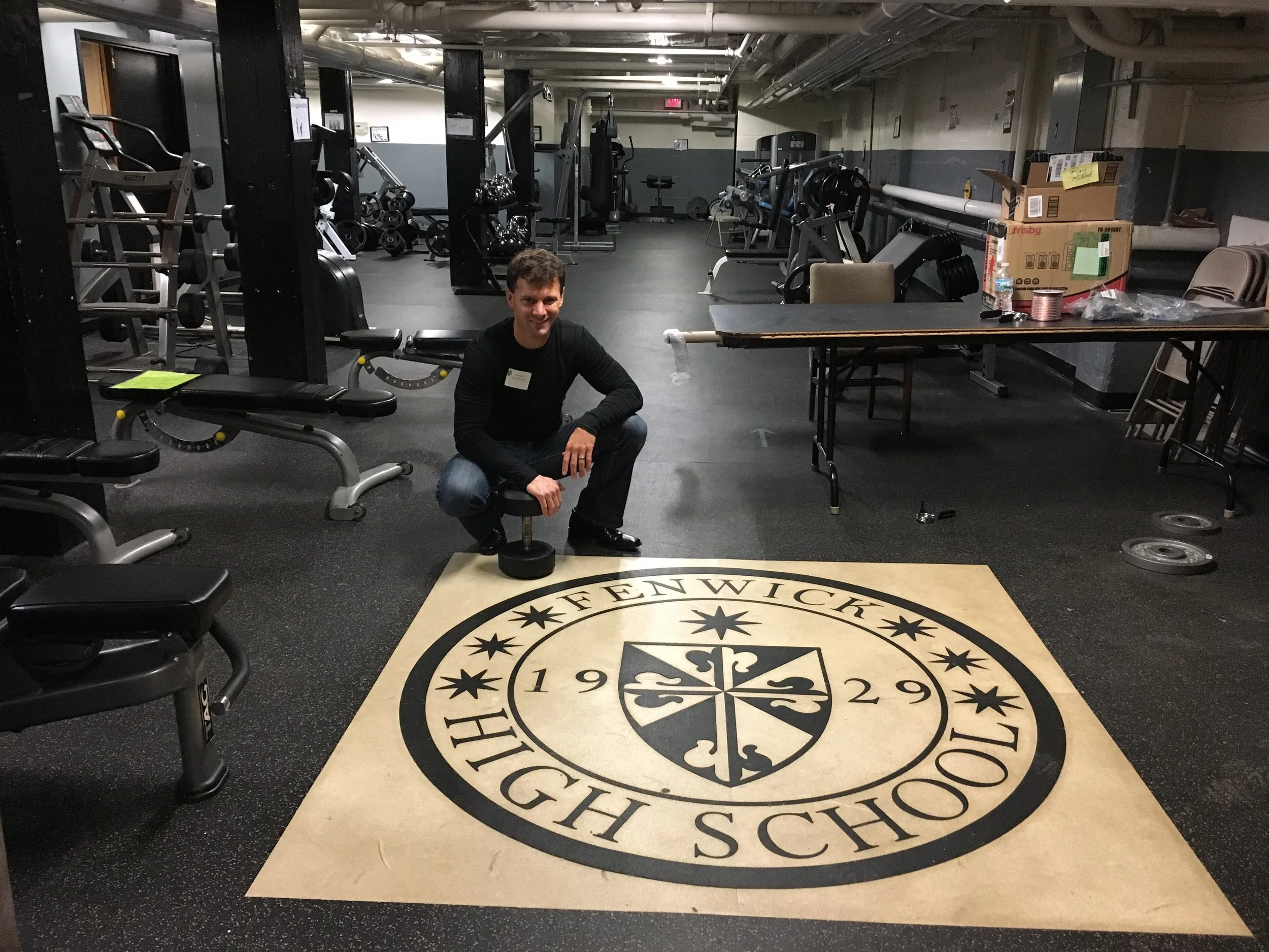 Michael Ockrim, founder of Mighty Oak Athletic in Westmont, Illinois, crouching beside a dumbbell on the Fenwick High School 1929 crest in the school’s basement weight room, where his first workout took place in 1992.