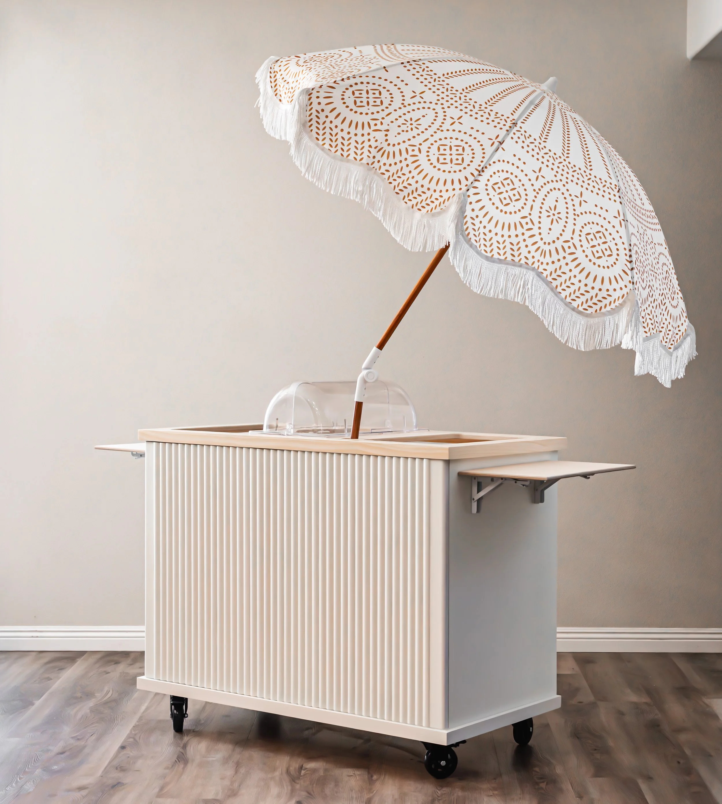 A white fluted charcuterie cart with a beige slatted front, four caster wheels, a large decorative umbrella with a white fringe and brown pattern on top, and a small transparent cover on the top surface.