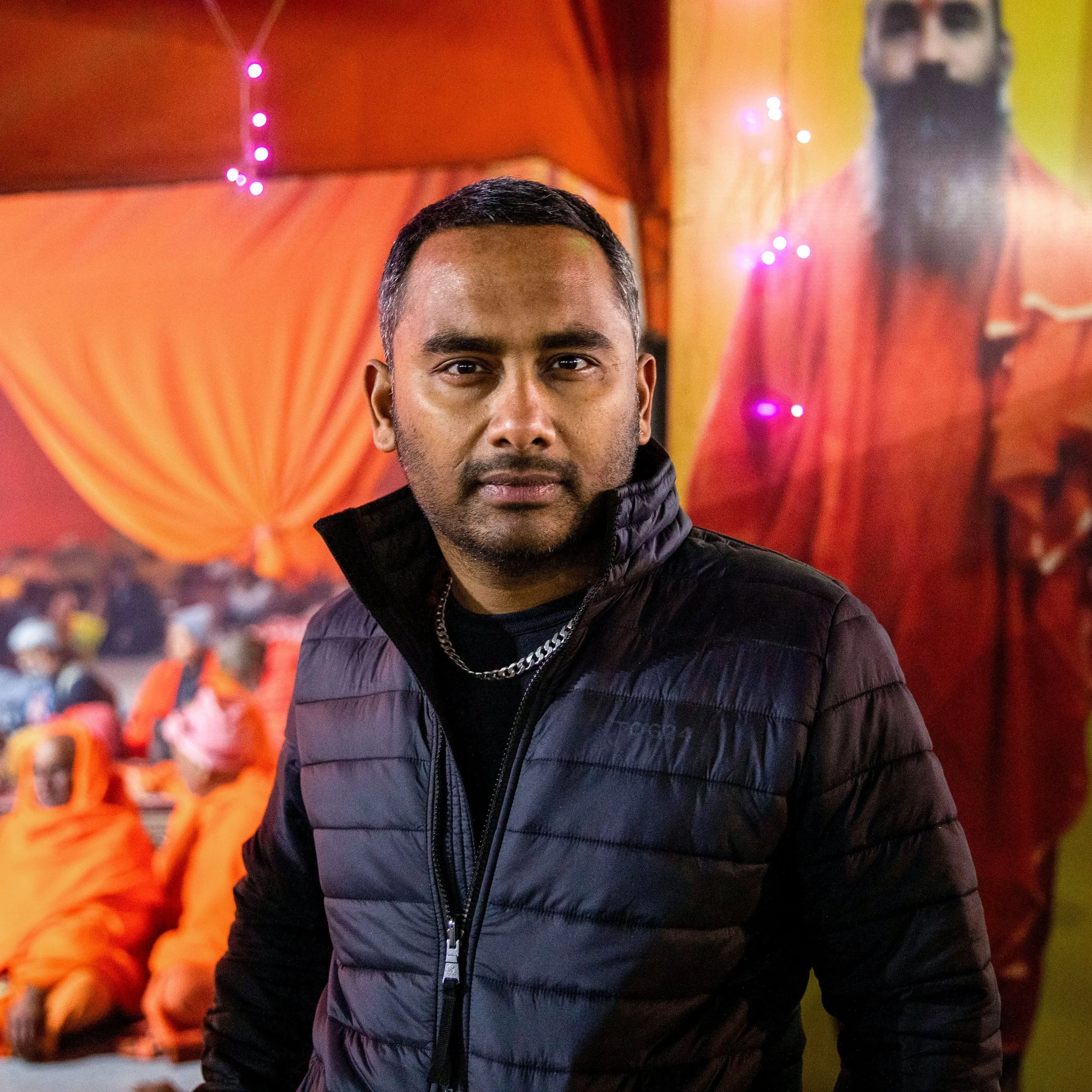 A man with short black hair, a mustache, and a goatee, wearing a black puffer jacket and a silver chain, stands in front of a vibrant orange and yellow backdrop with a large portrait of a man with a beard and traditional attire. Orange-clad monks are seated in the background.
