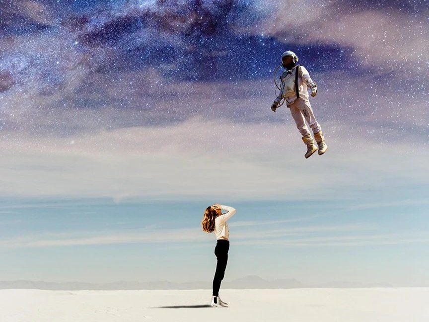 A woman standing on a barren landscape, looking up at an astronaut floating in the sky among stars and the Milky Way galaxy.