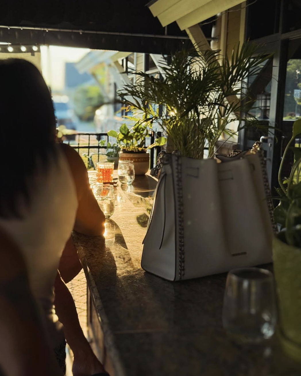 A woman sitting at a bar or counter during sunset, with sunlight casting shadows on her and illuminating a white handbag and drinks on the counter, decorated with potted plants.