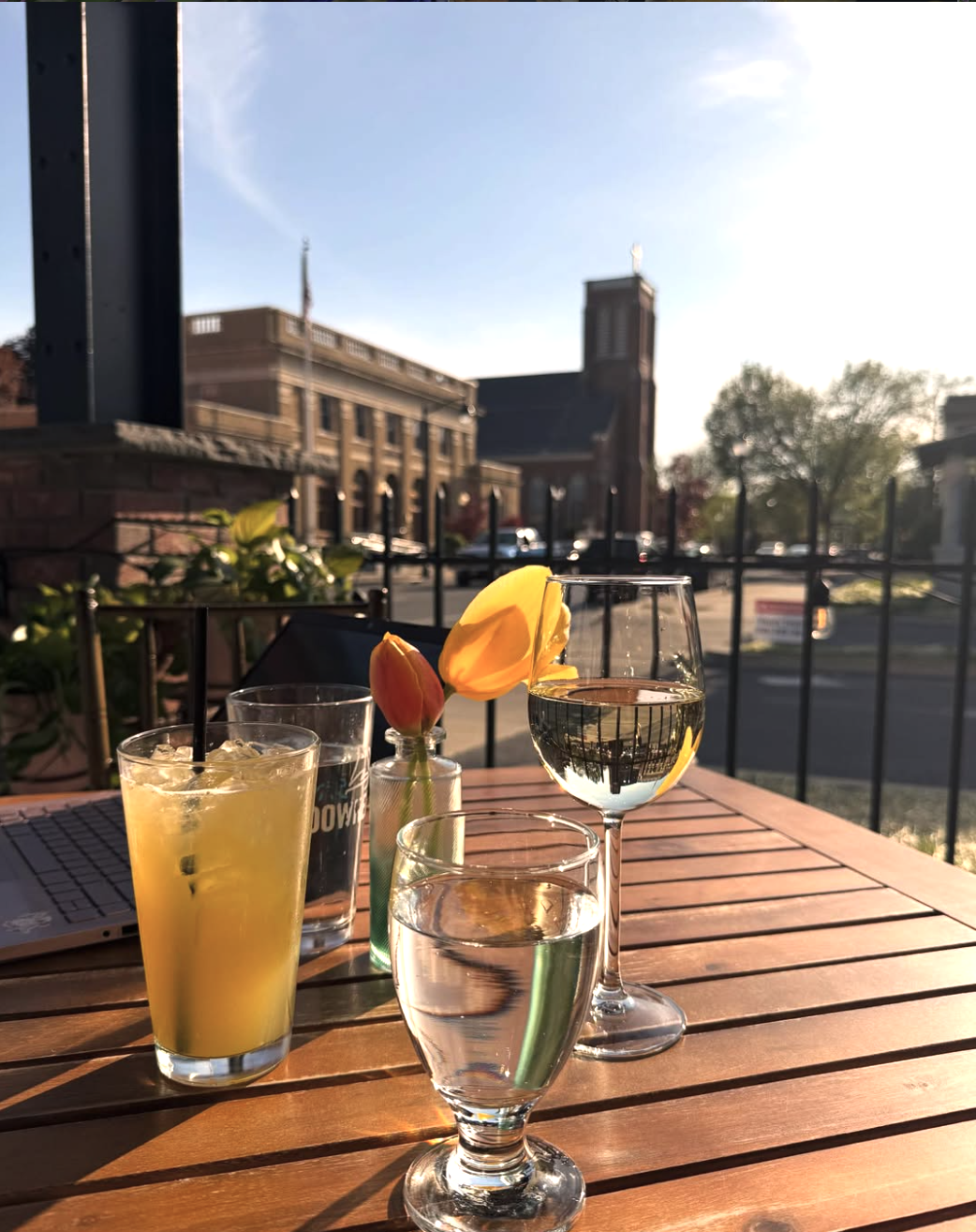 A wooden outdoor table with three drinks: a glass of orange juice, a glass of water, and a glass of white wine, with a tulip in a small vase, set against a cityscape background with a building and trees, during sunset.