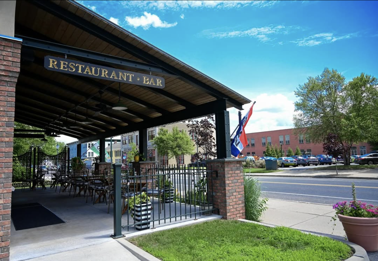 Exterior of a restaurant and bar with patio seating, potted plants, flag, and greenery, across from a parking lot and street, on a clear day.