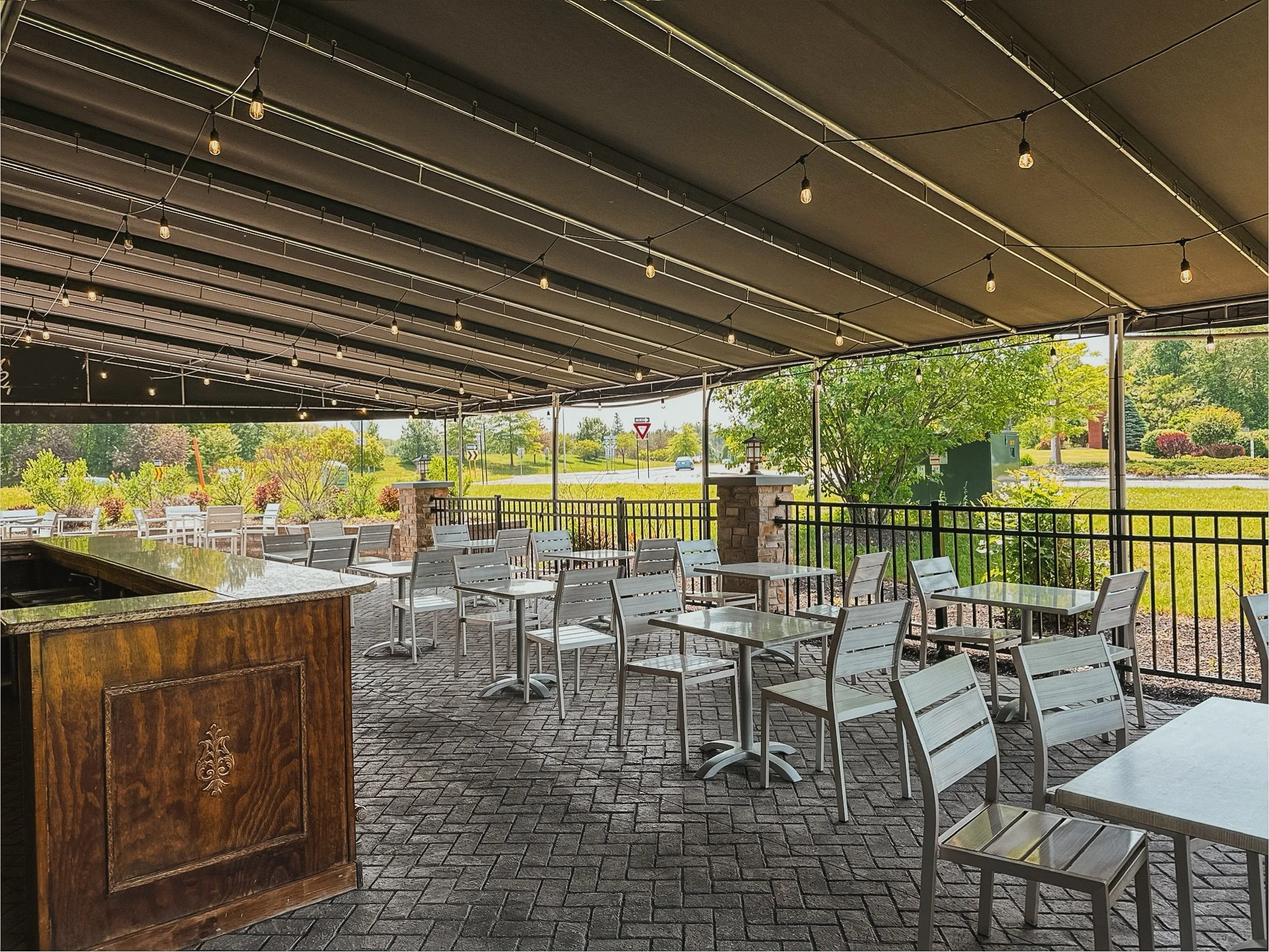 An outdoor patio covered with a black canopy and string lights, with metal tables and chairs, surrounded by greenery and trees.