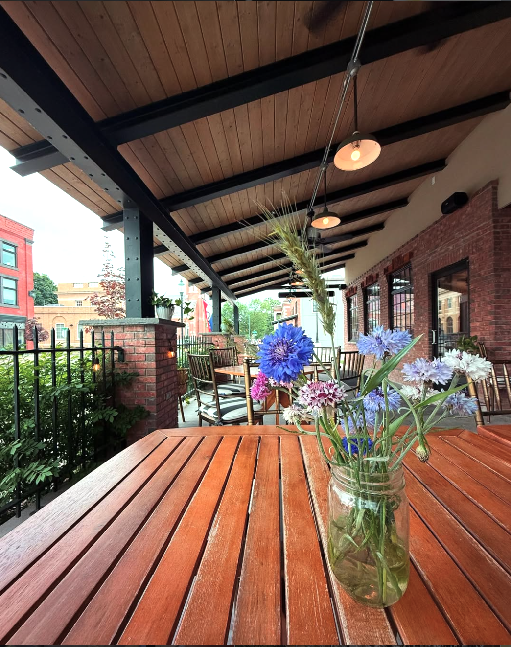 A cozy outdoor restaurant patio with wooden furniture, a glass jar with wildflowers on a table, brick walls, and string lights hanging from a wooden ceiling.