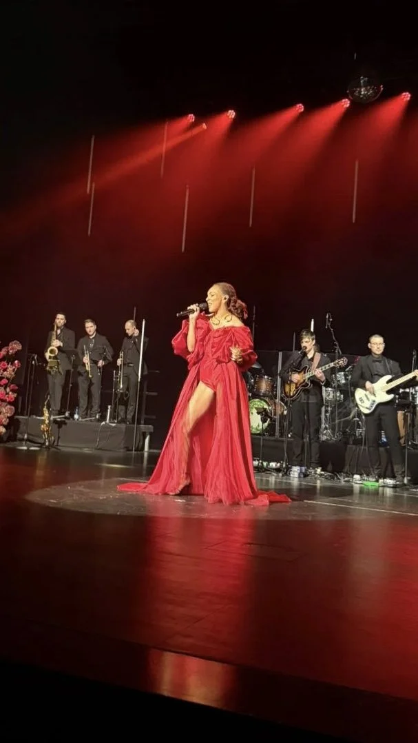 A woman in a glamorous red dress performing with a microphone on a stage, accompanied by a band with several musicians playing guitars, keyboards, and drums, under red stage lighting.