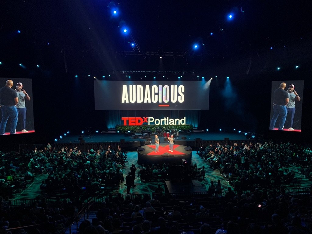 Large audience attending a TEDx event in Portland, with a stage featuring a large screen displaying the word 'AUDACIOUS' and two speakers on stage, flanked by side screens showing one of the speakers.