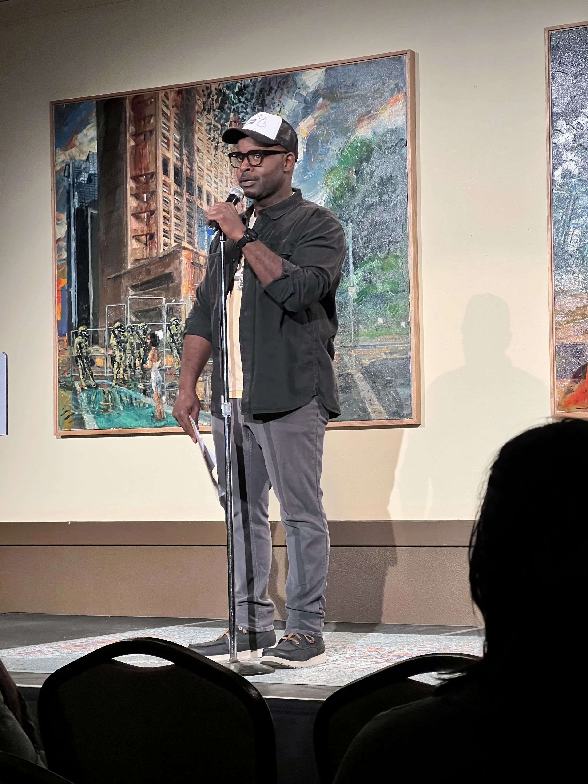 Chris with glasses, wearing a cap and casual clothes, speaking into a microphone on a stage in front of paintings, with an audience seated and visible in the foreground.
