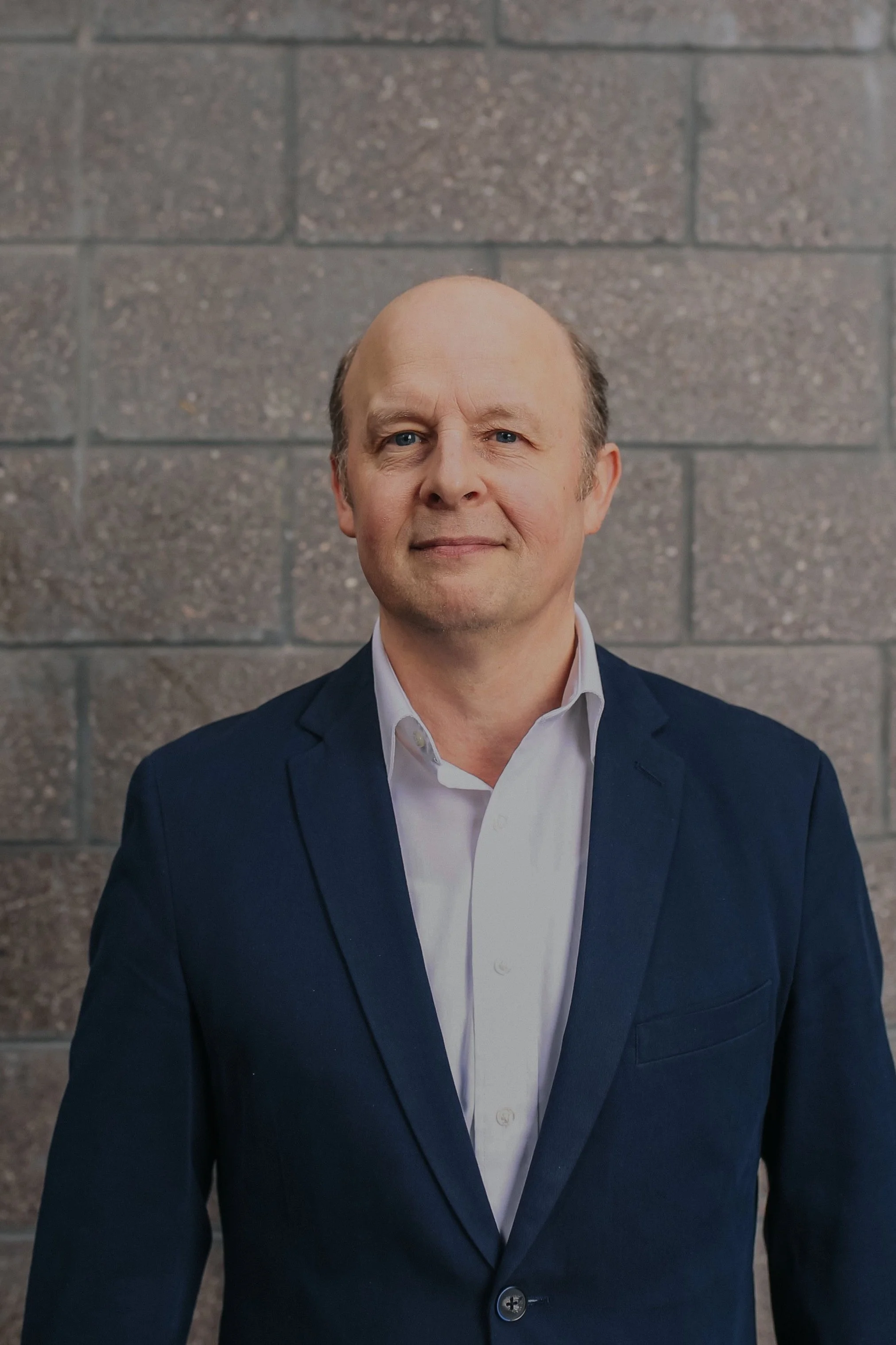 Headshot of a man in a suit against a brick wall background.