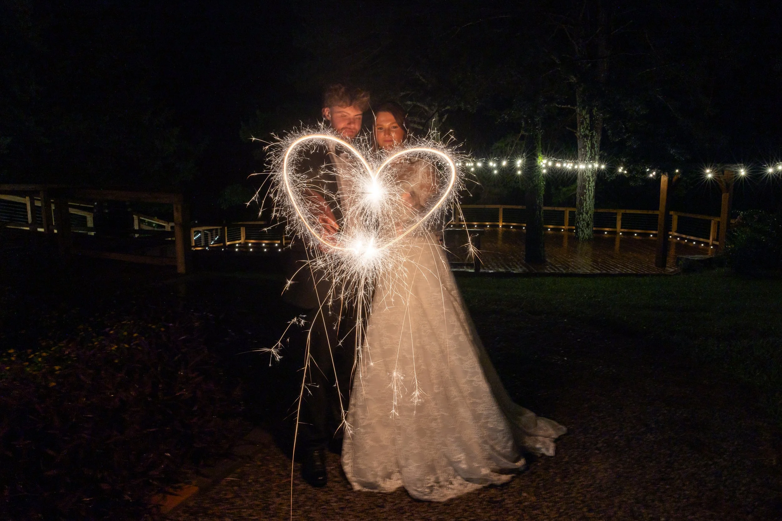A heart is being drawn with a sparkler in a night-time wedding portrait.
