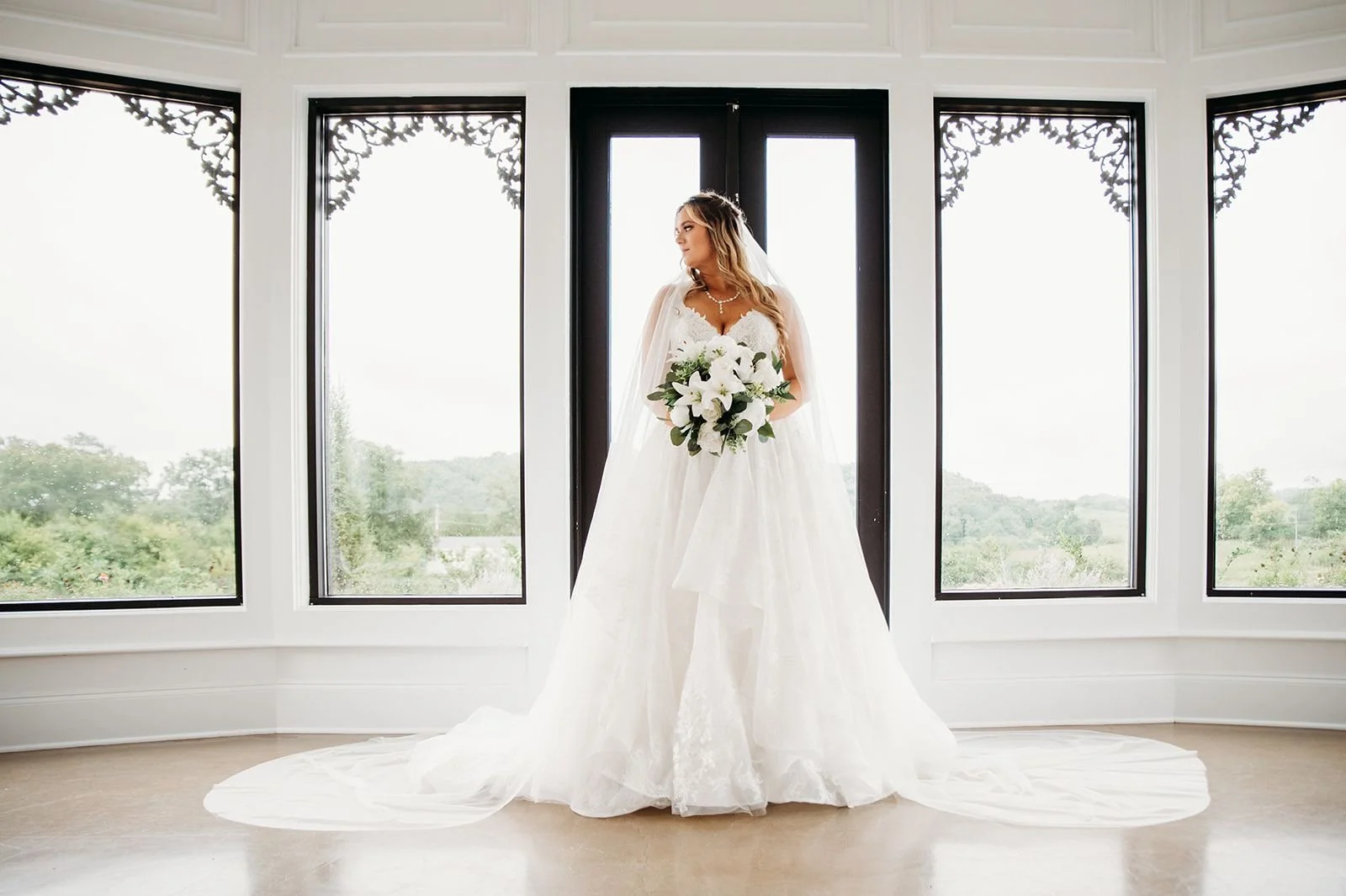 A bride poses indoors by herself in front of large windows.