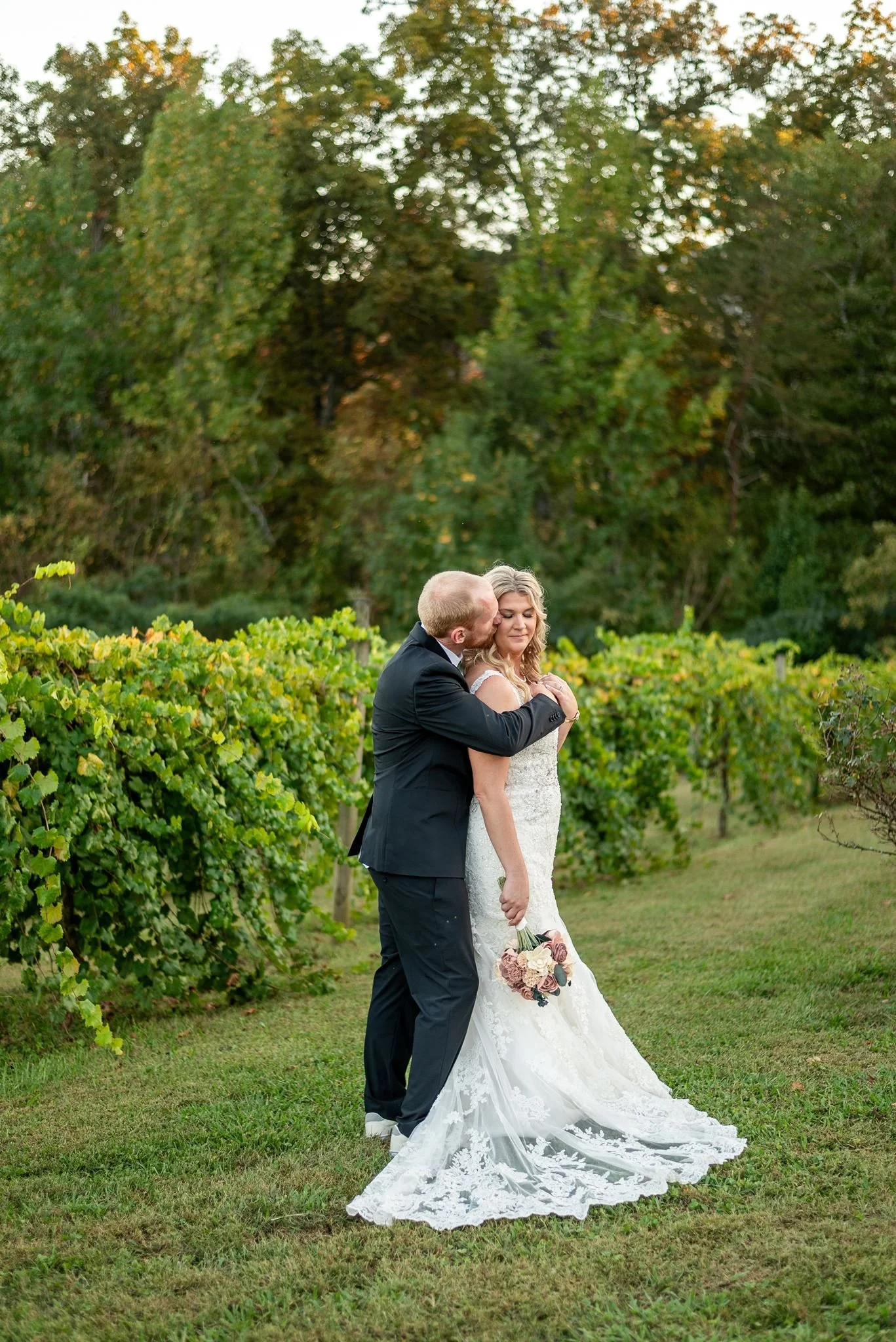 The wedding couple poses with the groom standing behind the bride in the middle of the vineyard.