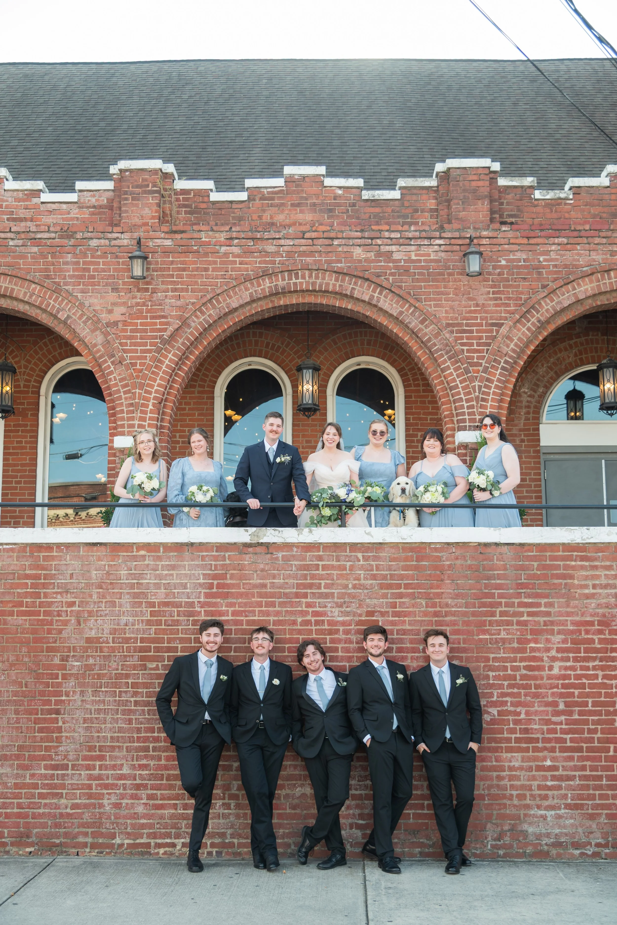 A bridal party poses outside of their brick venue with girls on top and boys on bottom.