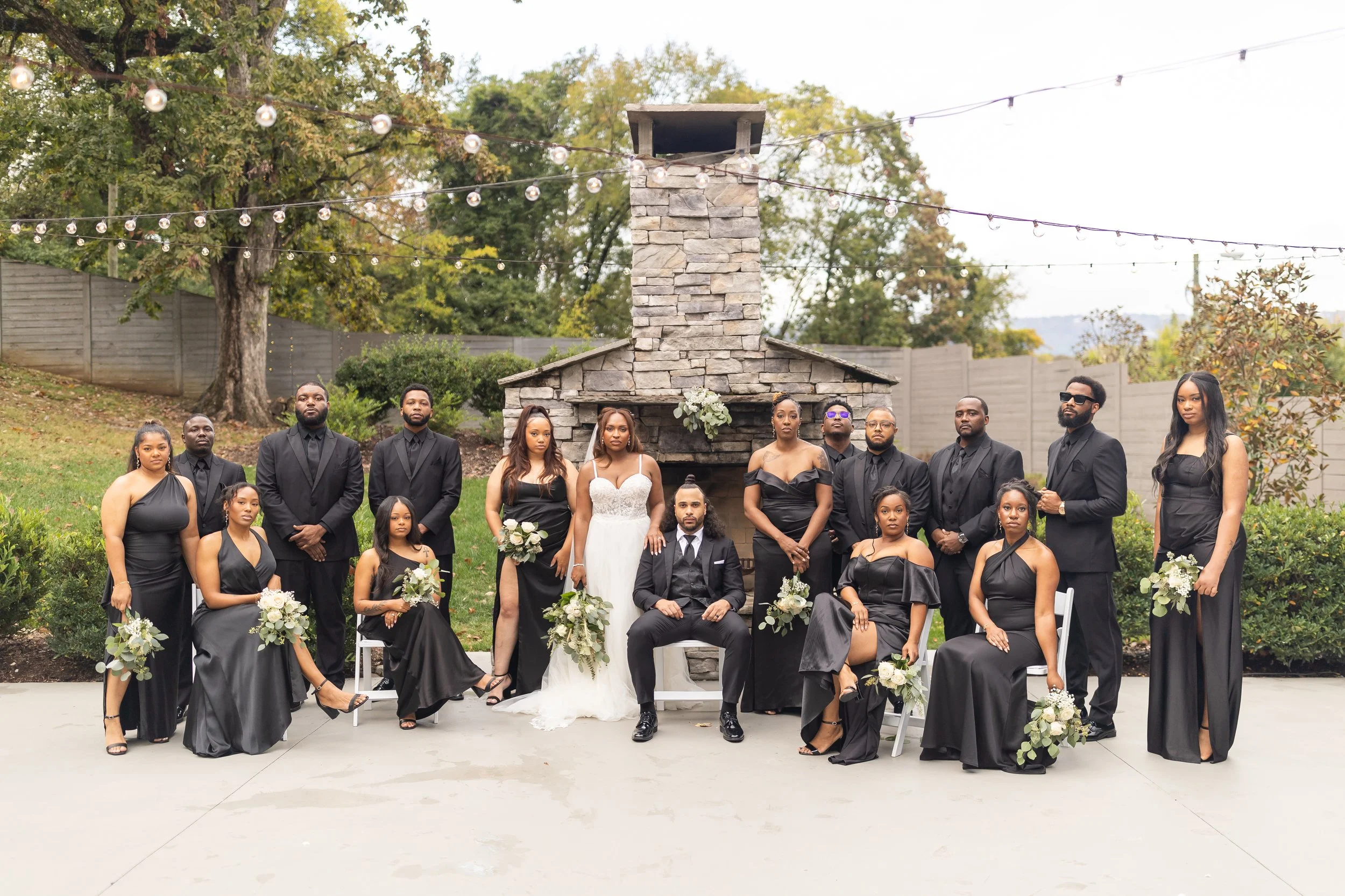 A large bridal party stands in front of a fireplace in all black.