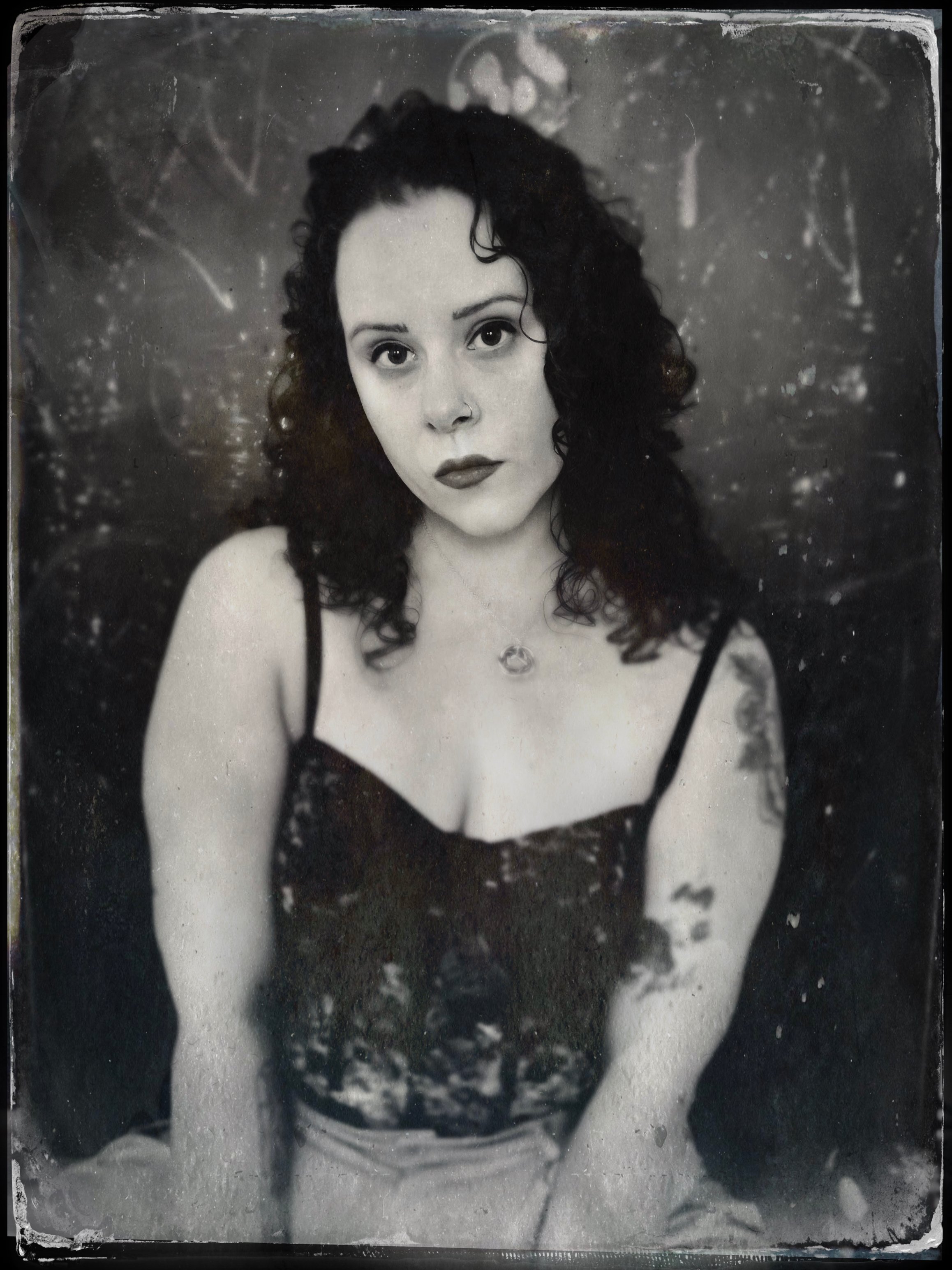 A black and white portrait of a woman with dark, curly hair, wearing a lace top and a necklace, against a textured background.