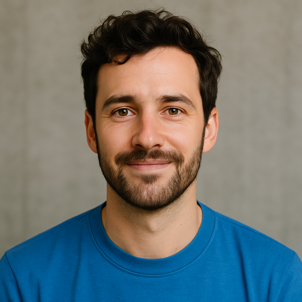 A man with dark, curly hair and a beard, wearing a blue shirt, smiling at the camera against a plain, light-colored background.