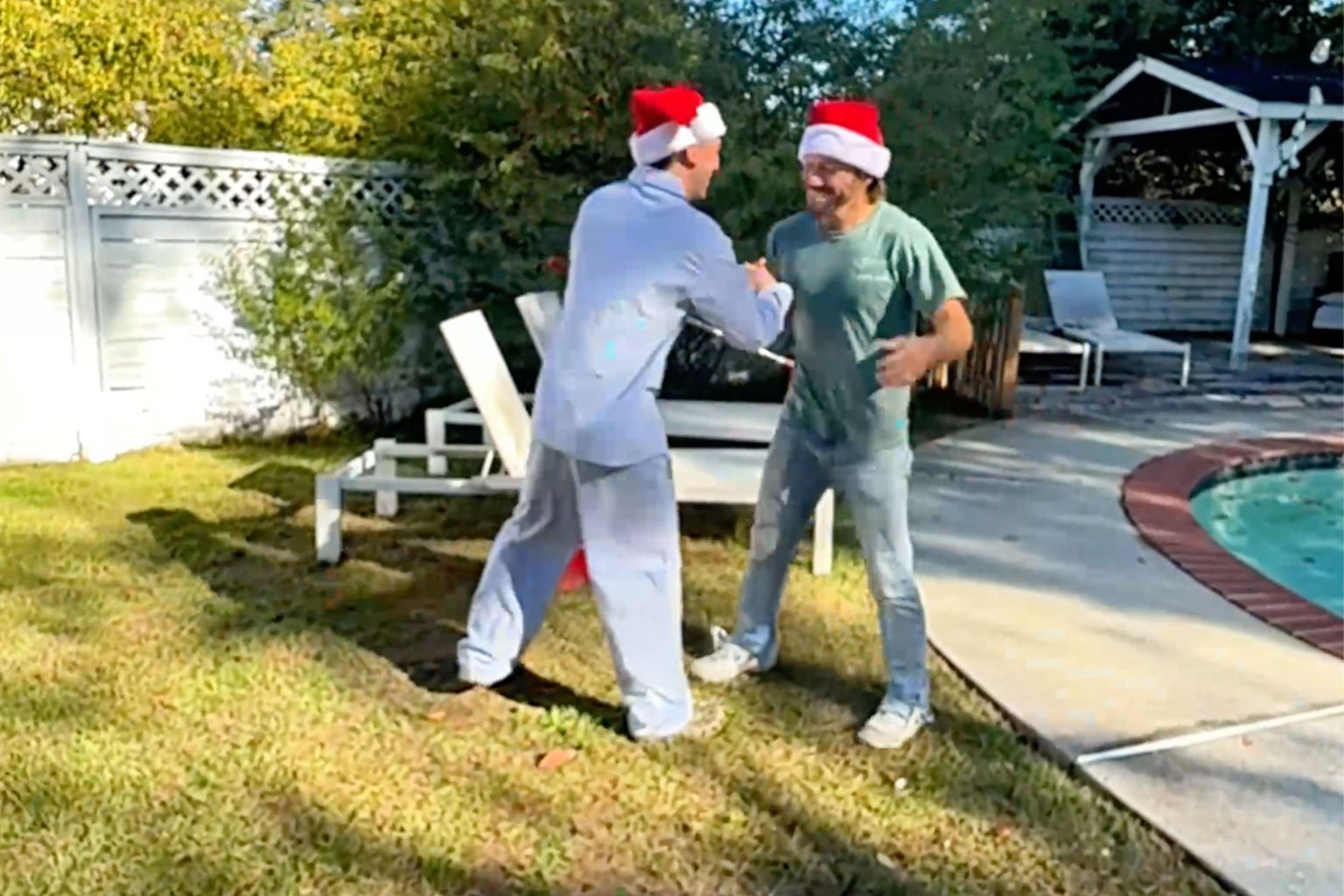 Two social media influencers wearing Santa hats exchanging a fist bump near a backyard pool, with poolside chairs and a gazebo visible.