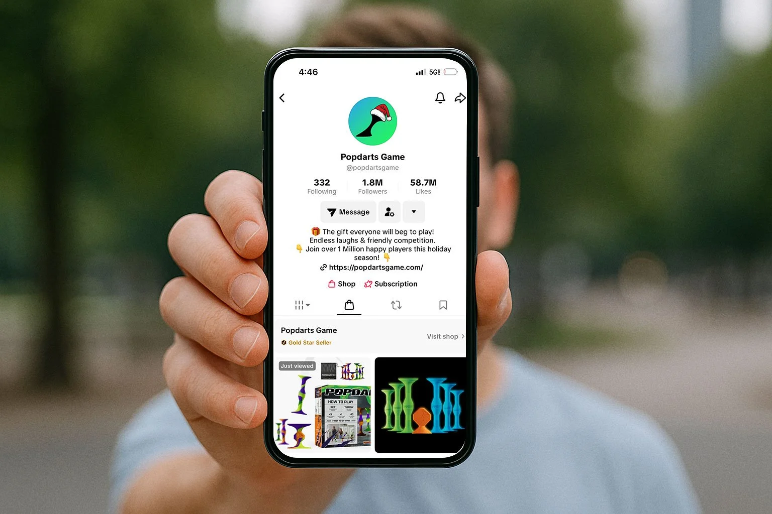 A person holding a smartphone with a TikTok profile named Popdarts Game visible on the screen. The profile displays a logo of a black dart with a red Santa hat, along with follower and like counts, and a bio about playing holiday dart games.
