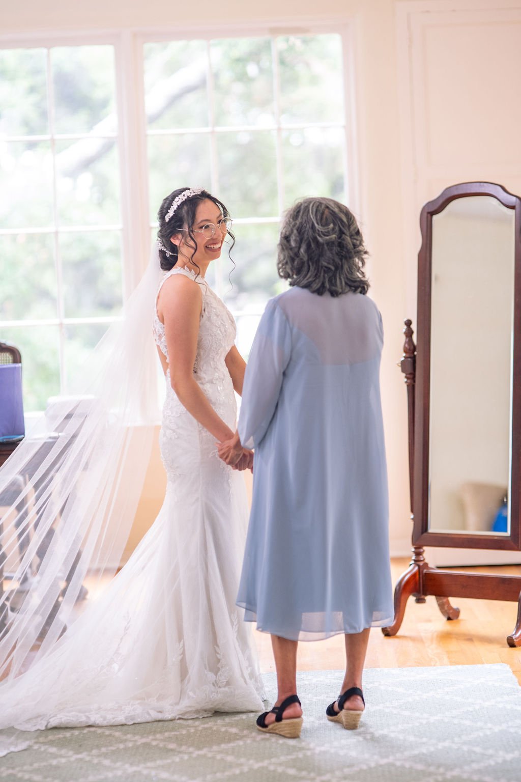 Bride in wedding dress holding hands with older woman in a light blue outfit in a bright room, standing near a mirror.