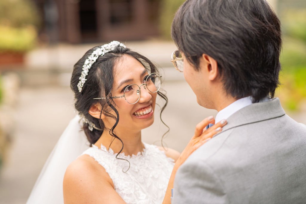 A bride wearing a white dress and glasses smiles at her partner, who is dressed in a light gray suit, outdoors.