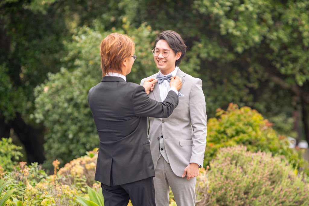Two men outdoors, one adjusting the other's bow tie, both smiling, with greenery in the background.
