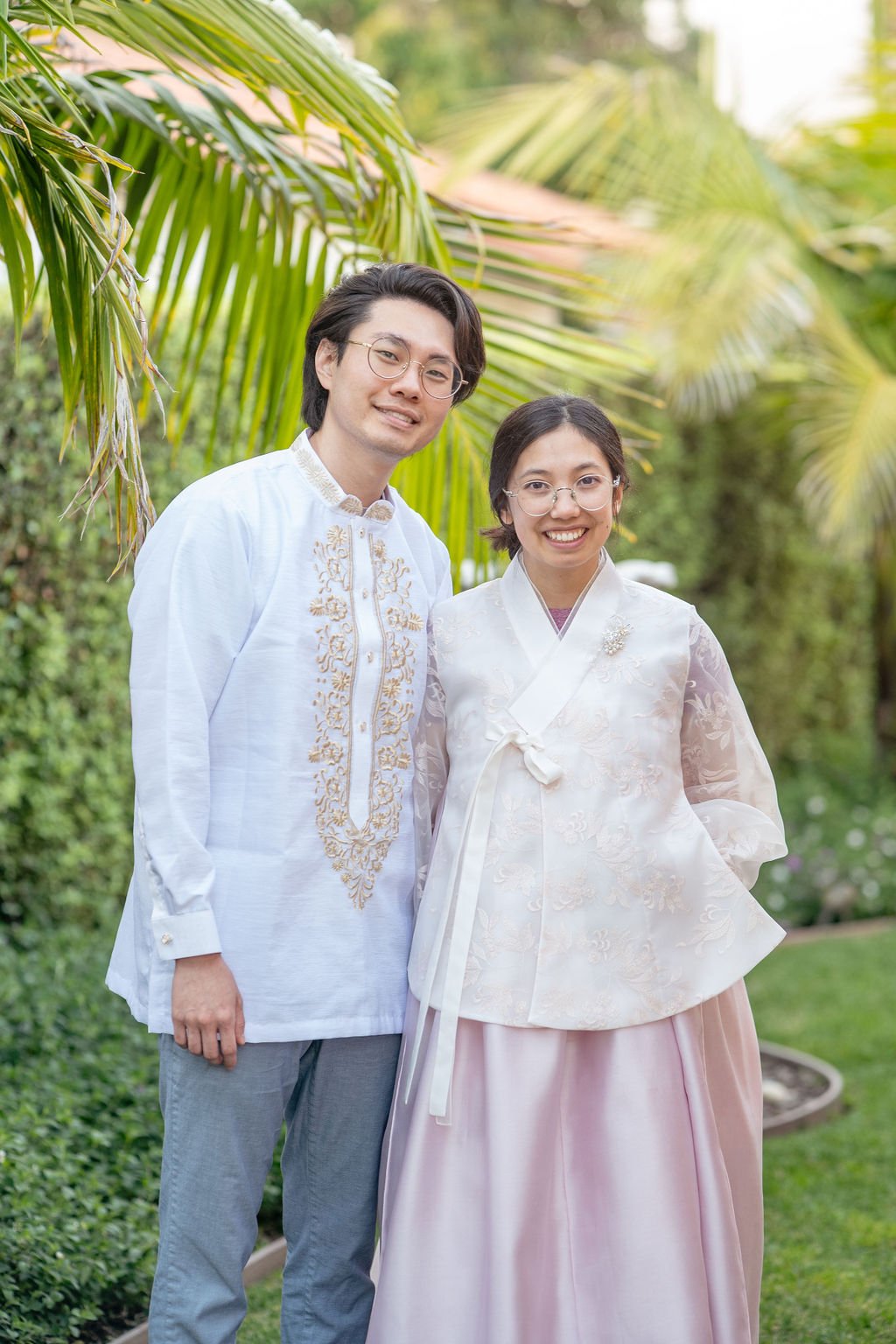 A couple in traditional attire standing outside next to green foliage, smiling at the camera.