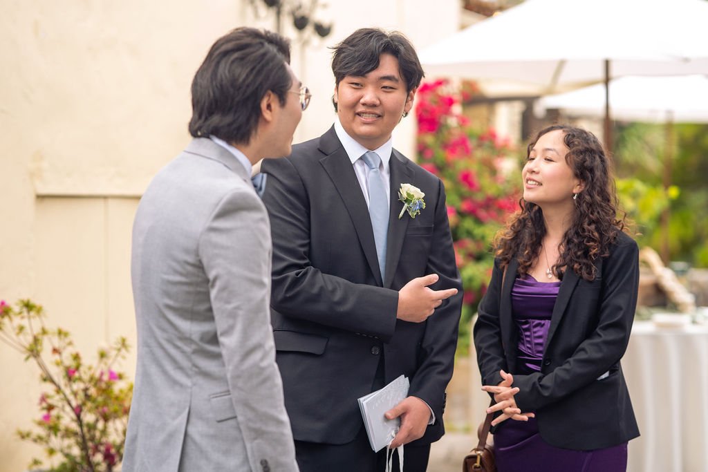 Three people in formal attire conversing outdoors at an event, with flowers and a table in the background.