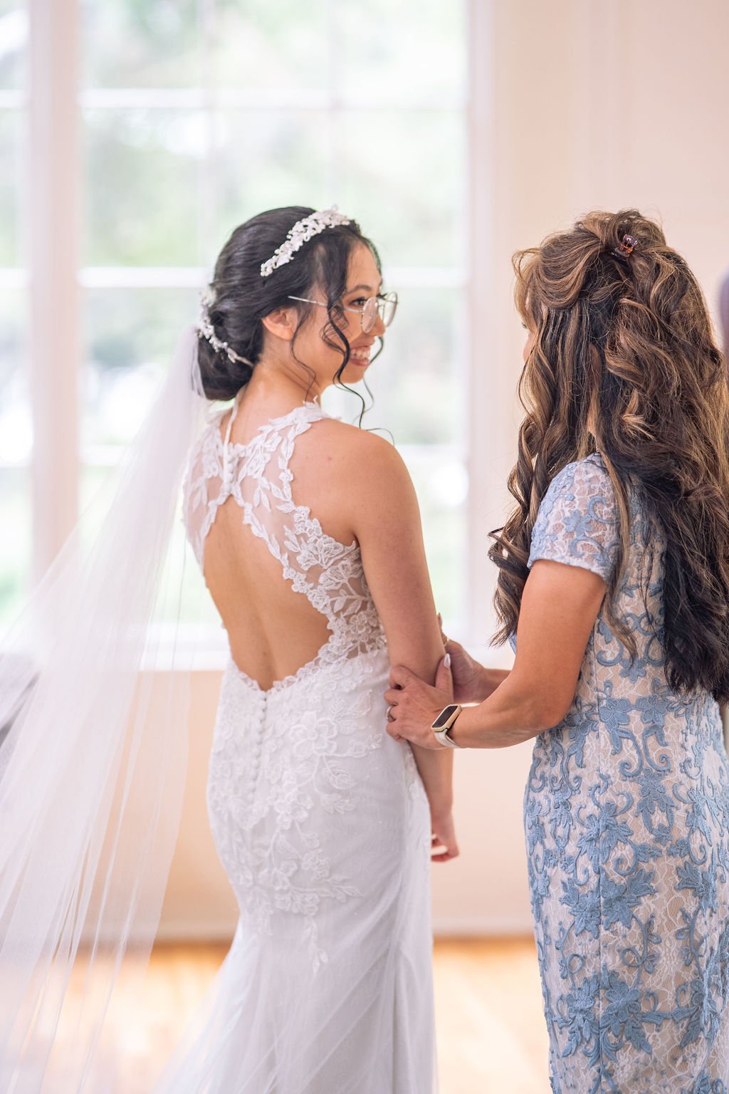 Bride in lace wedding dress and veil, standing with a woman in a blue patterned dress, indoors.