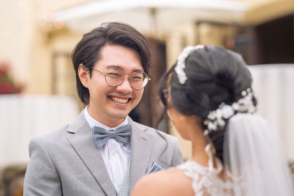 Bride and groom smiling at each other during a wedding ceremony, with the groom wearing a gray suit and glasses, and the bride adorned with a veil and hair accessories.