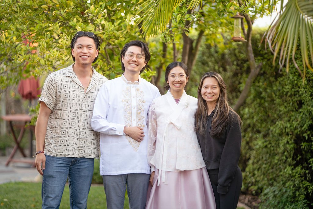 Group photo of four people outside, two wearing traditional Asian attire, and two in casual clothing, standing in a garden with green foliage.