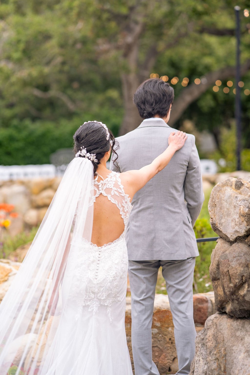 Bride in lace wedding dress gently touching groom in gray suit from behind, outdoors with greenery.