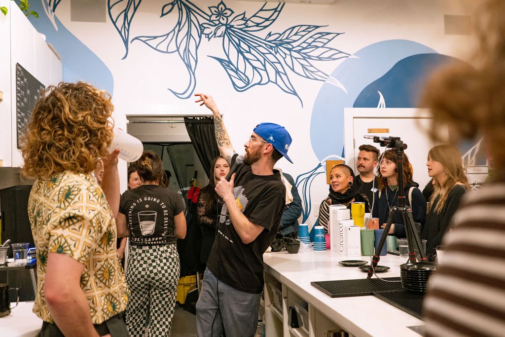 A group of people in a contemporary coffee shop, some listening attentively and one person gesturing energetically. The backdrop includes a mural with floral and abstract designs. Several cups and a camera on a tripod are visible on the counter.