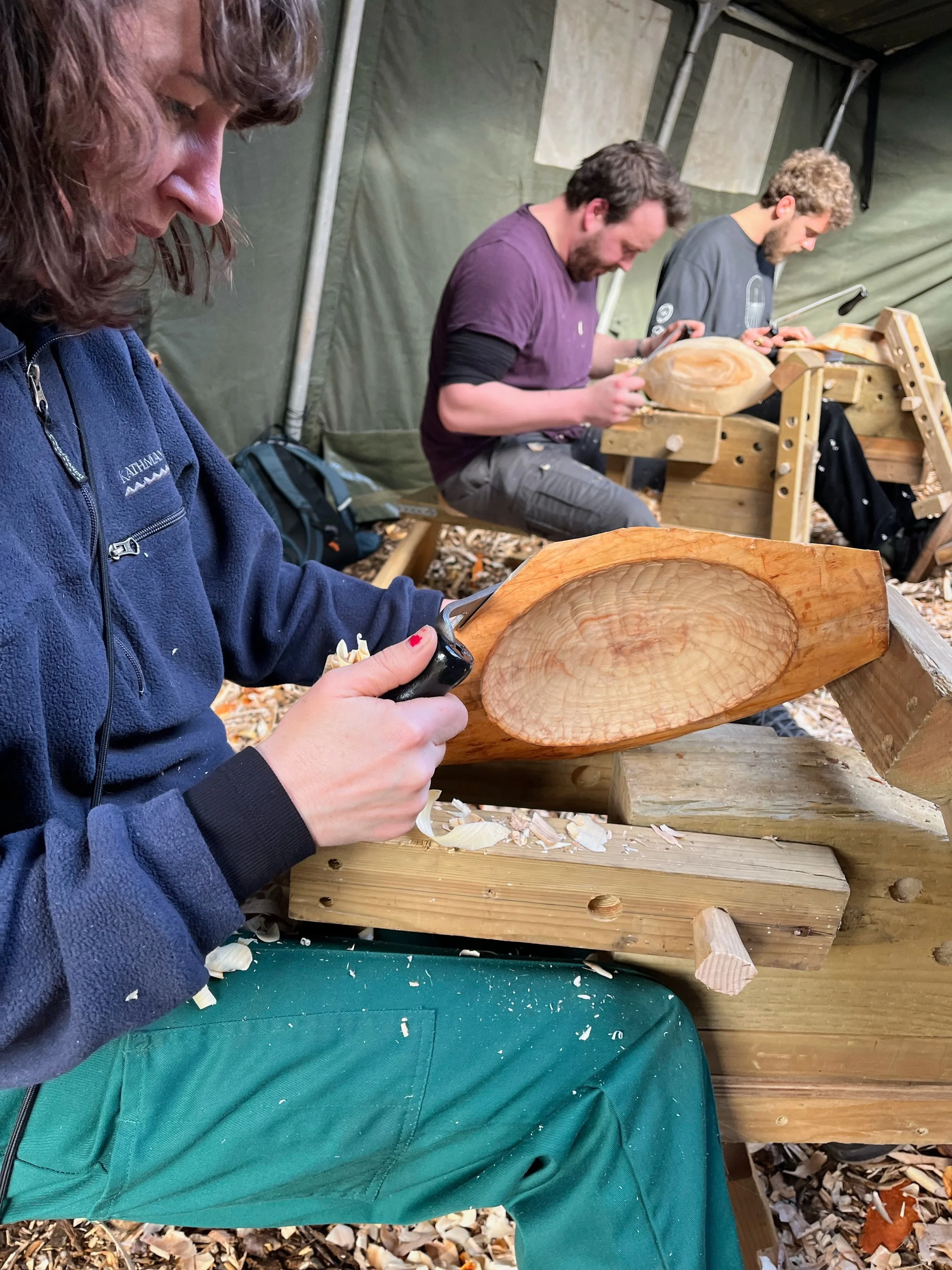 Three people carving wood on a woodworking bench inside a tent, with wood shavings on the ground.