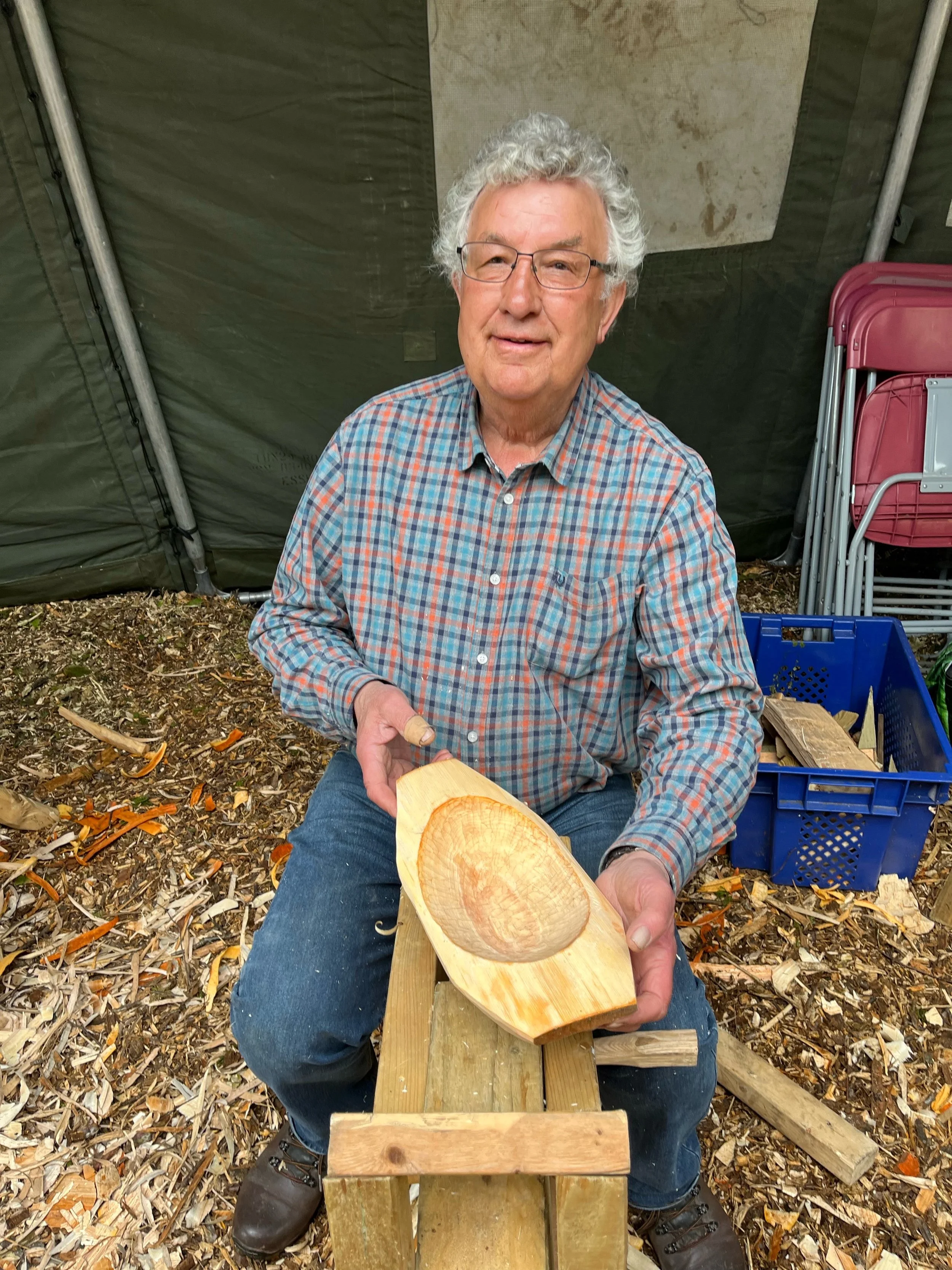 An elderly man with gray hair, glasses, and a plaid shirt is sitting outdoors, holding a carved wooden bowl or spoon. He is surrounded by wood shavings, woodwork, and some stacked chairs and boxes behind him.