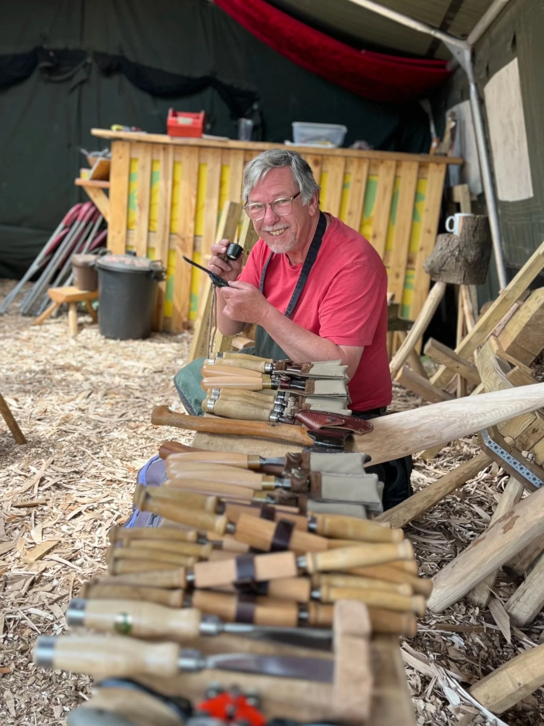 A man with glasses and a red shirt smiling while holding a camera in a woodworking shop. The table in front of him is filled with various woodworking tools, including chisels and carving knives. Wood shavings cover the floor, and a workbench and wood