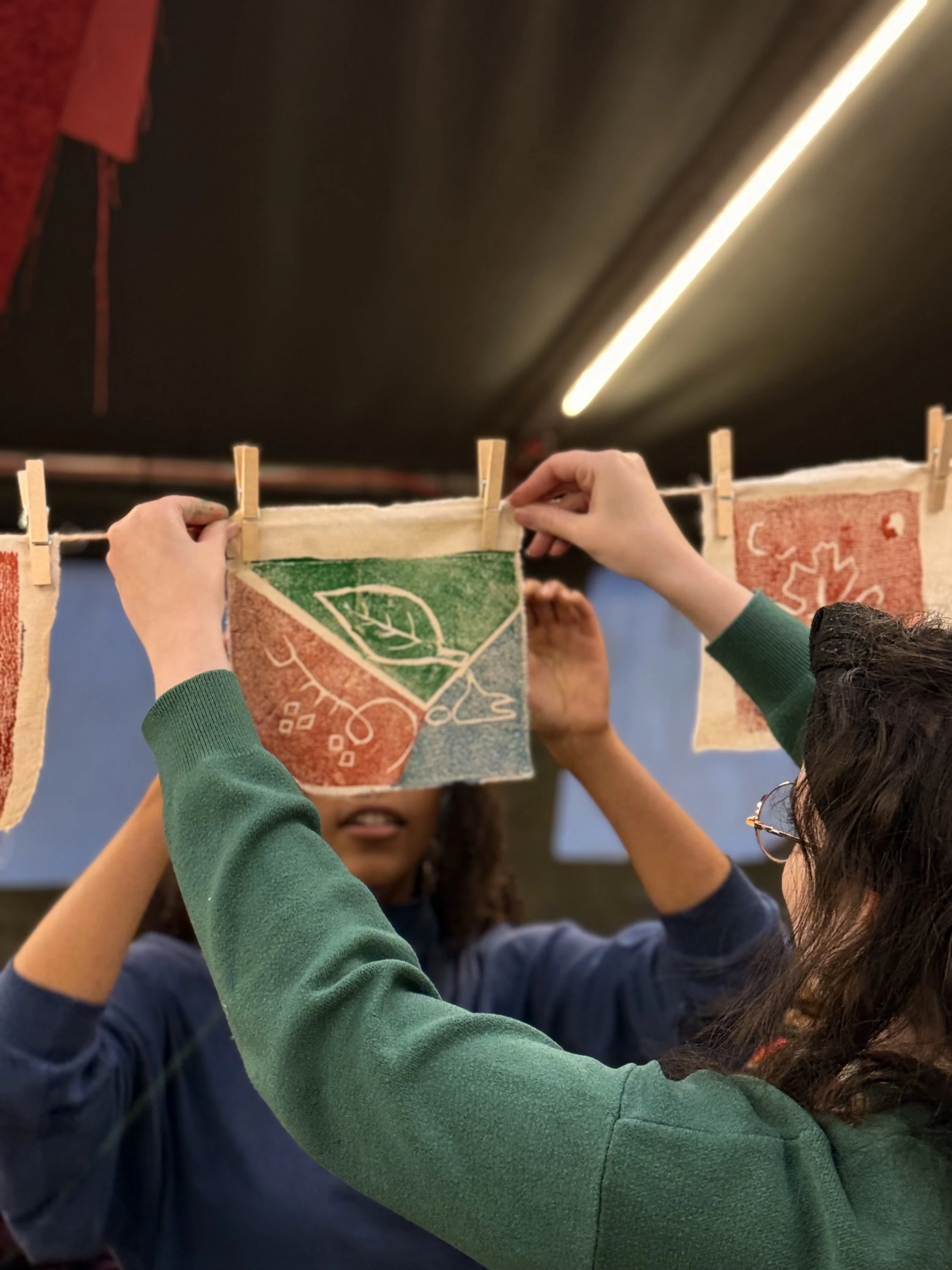 Two people hanging decorative fabric pieces on a clothesline inside a room.