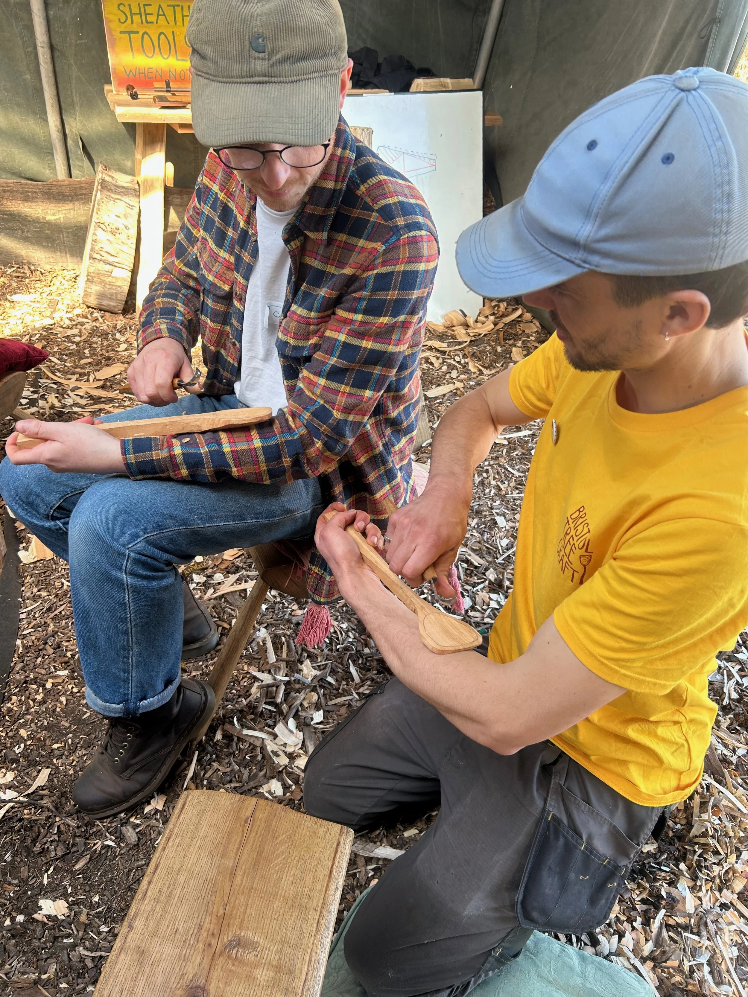 Two men, one wearing a plaid shirt and the other a yellow t-shirt, are working together on a woodworking project, shaping a piece of wood with hand tools inside a workshop with wood shavings on the ground.
