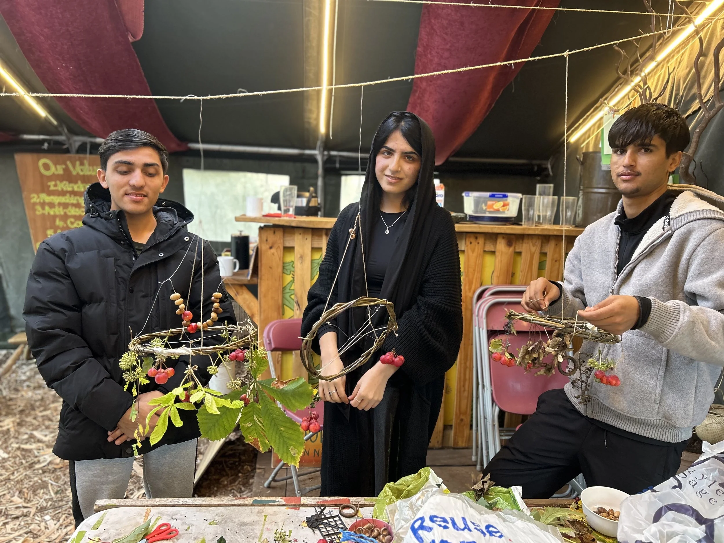 Three young people displaying handmade nature crafts with berries, leaves, and twigs inside a tent or outdoor space.