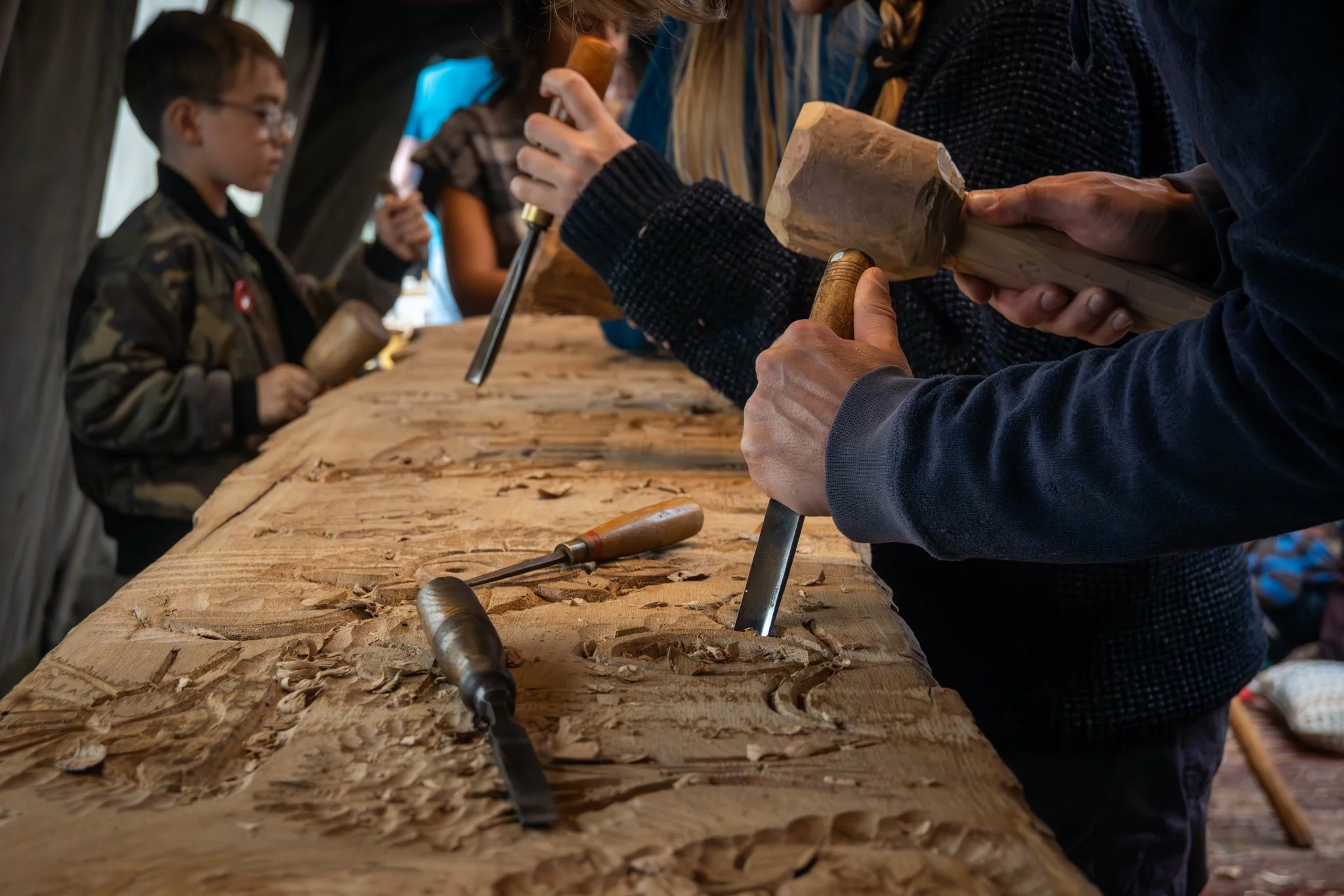 People working on wood carving at a workshop. Multiple chisels and carving tools are placed on a large wooden surface. A person in the foreground is holding a hammer and a chisel, while others are working on their carvings.
