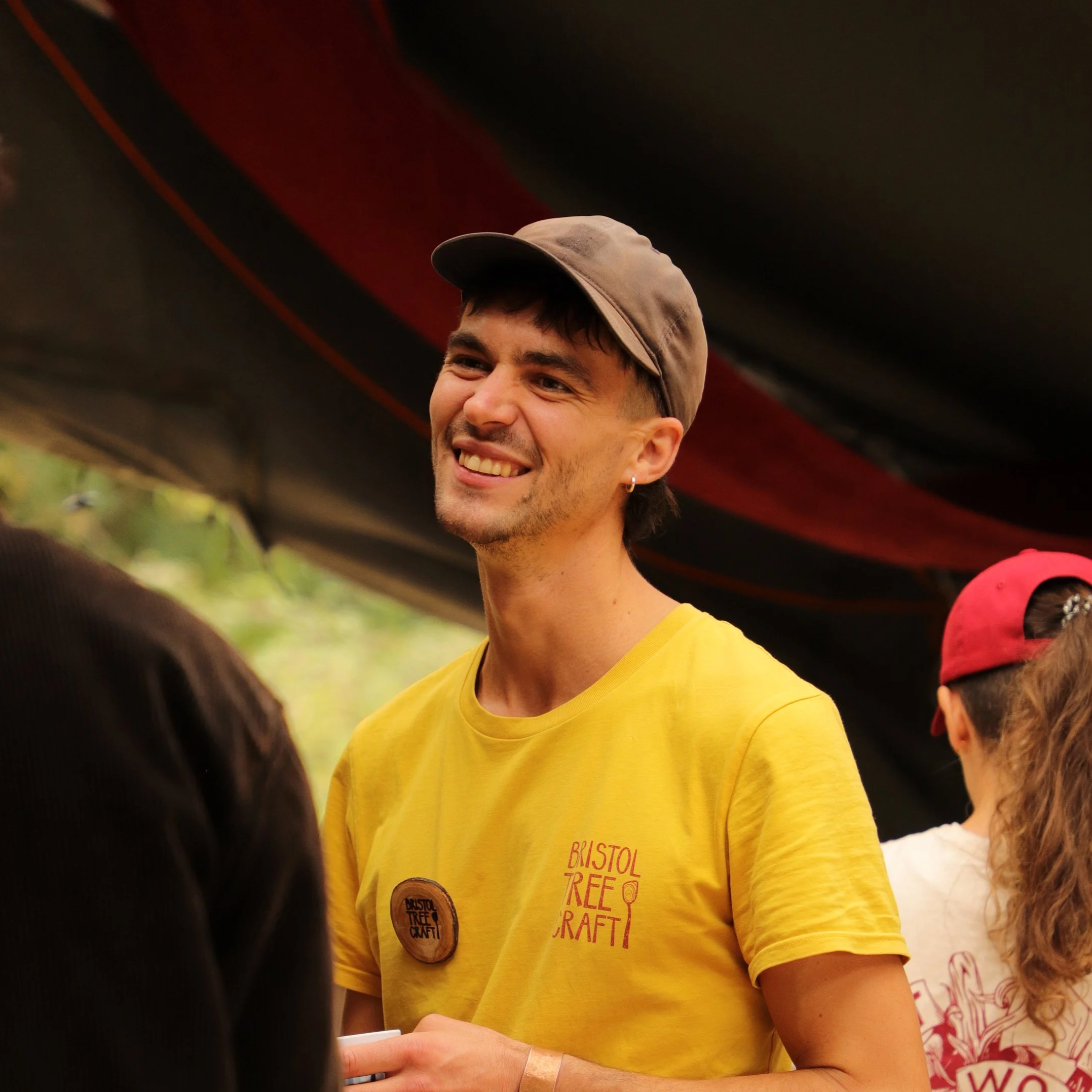 A young man with short dark hair, a beard, and a big smile, wearing a yellow T-shirt with 'Bristol Tree Craft' written on it, standing outdoors on a cloudy day with trees in the background.