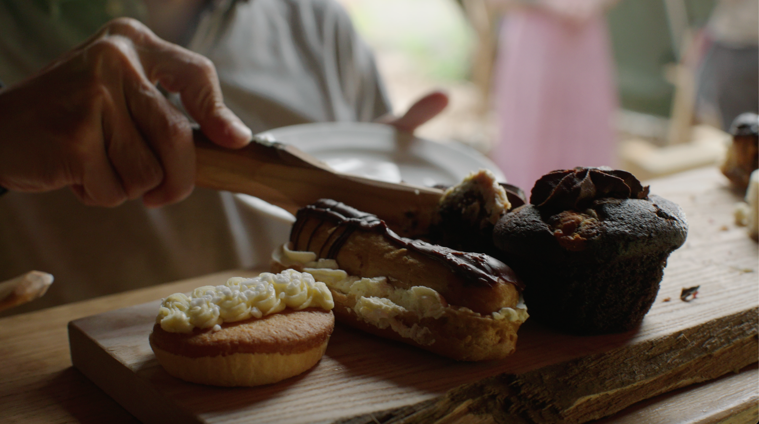 Hand holding a plate next to desserts, including a chocolate cupcake, an éclair, and a cream-topped tart, on a wooden tray.