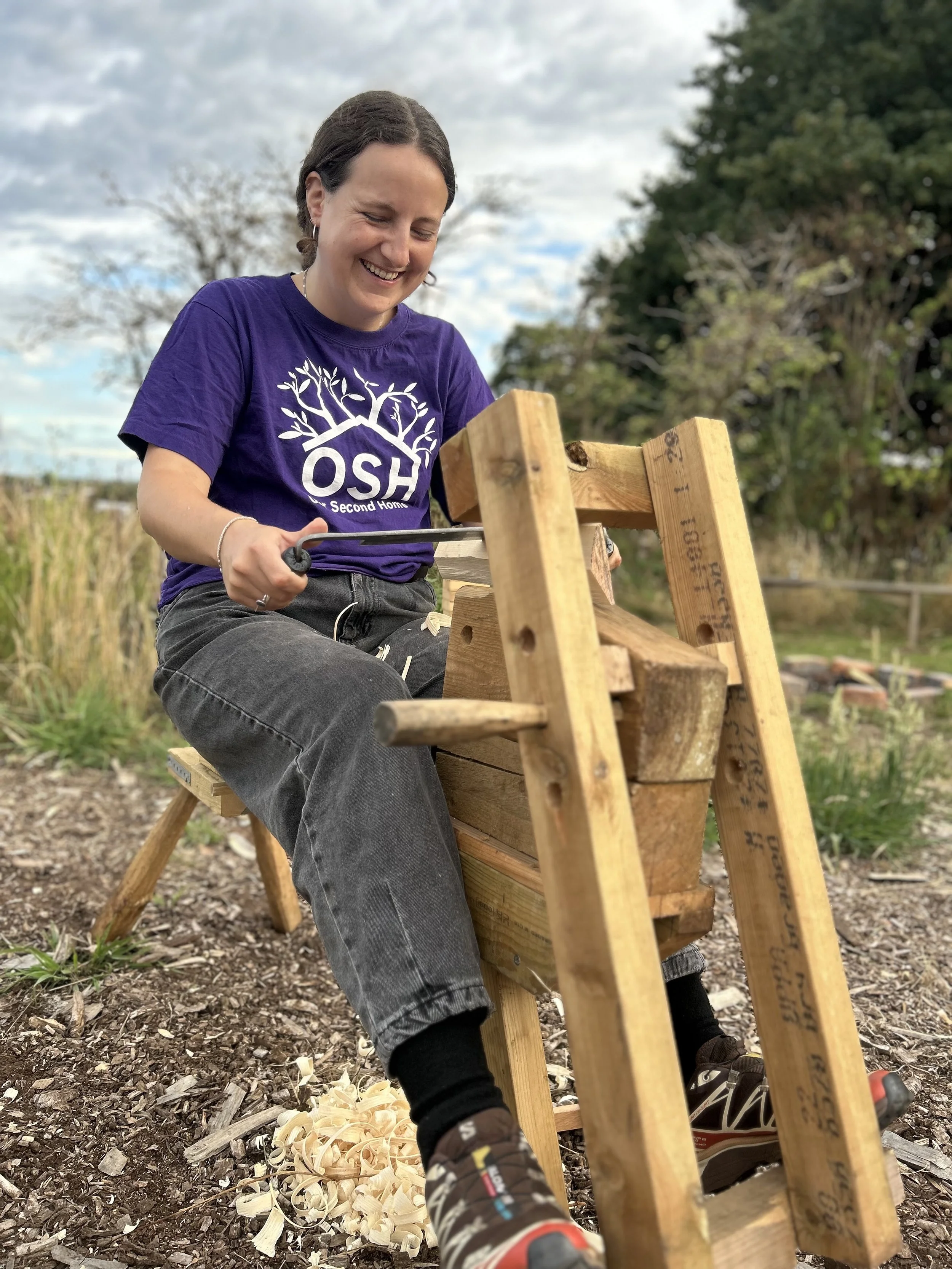 A woman in a purple t-shirt and gray jeans is sitting on a small wooden stool outside, carving or shaping wood on a sawhorse. She is smiling and focused on her work, with wood shavings on the ground nearby, surrounded by trees and a partly cloudy sky