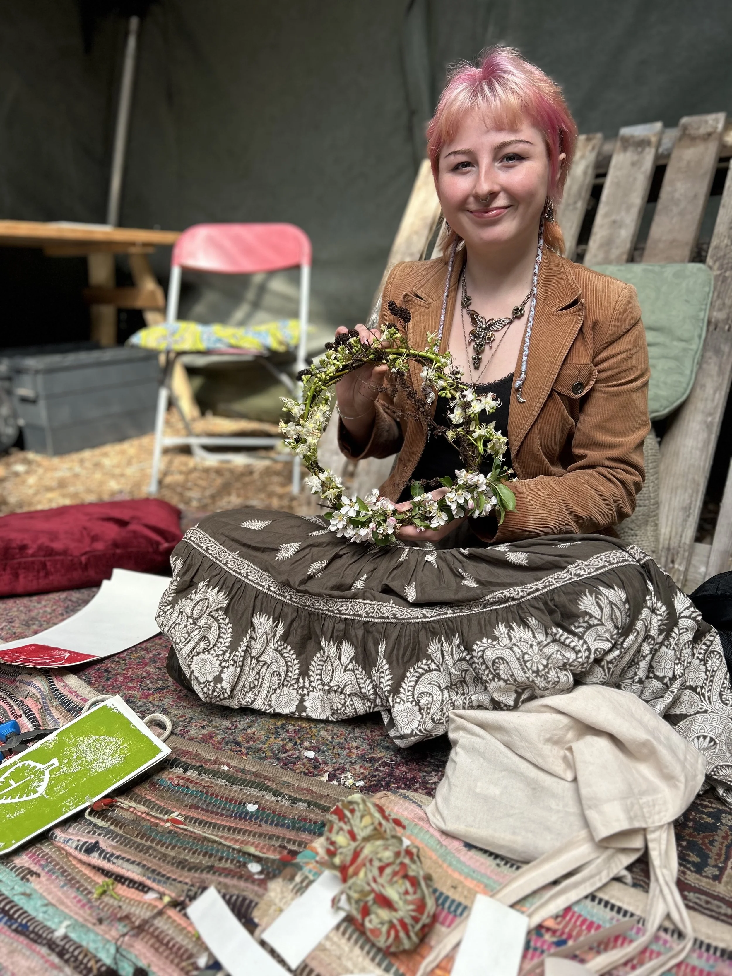 A woman with pink hair and a nose piercing smiling while holding a flower crown made of white flowers and green leaves. She is sitting on a colorful woven rug with sewing supplies and fabric in front of her, in what appears to be a crafting or outdoo