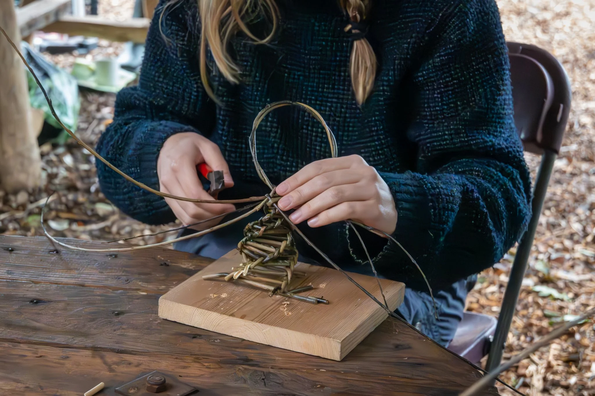 A person weaving a basket with natural materials at a wooden table outdoors, surrounded by fallen leaves.