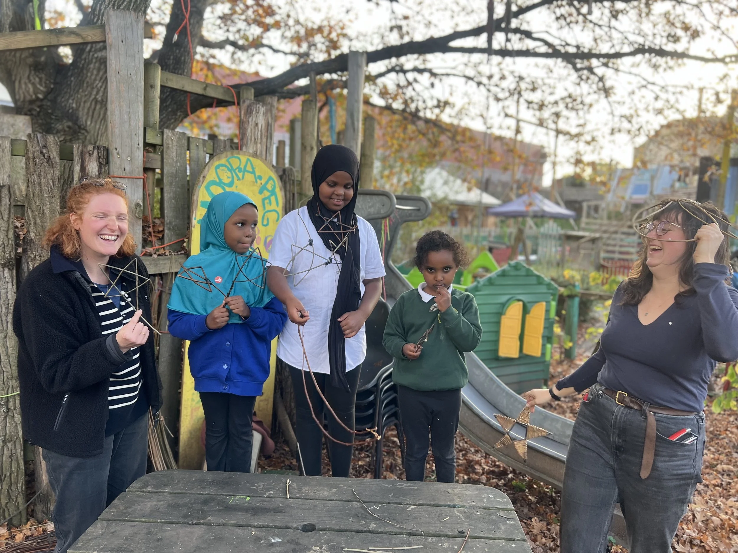 Five women and girls are outdoors in a park, holding star-shaped craft made of sticks. They are smiling and laughing, dressed in casual clothes, with a wooden fence and colorful playhouses in the background.