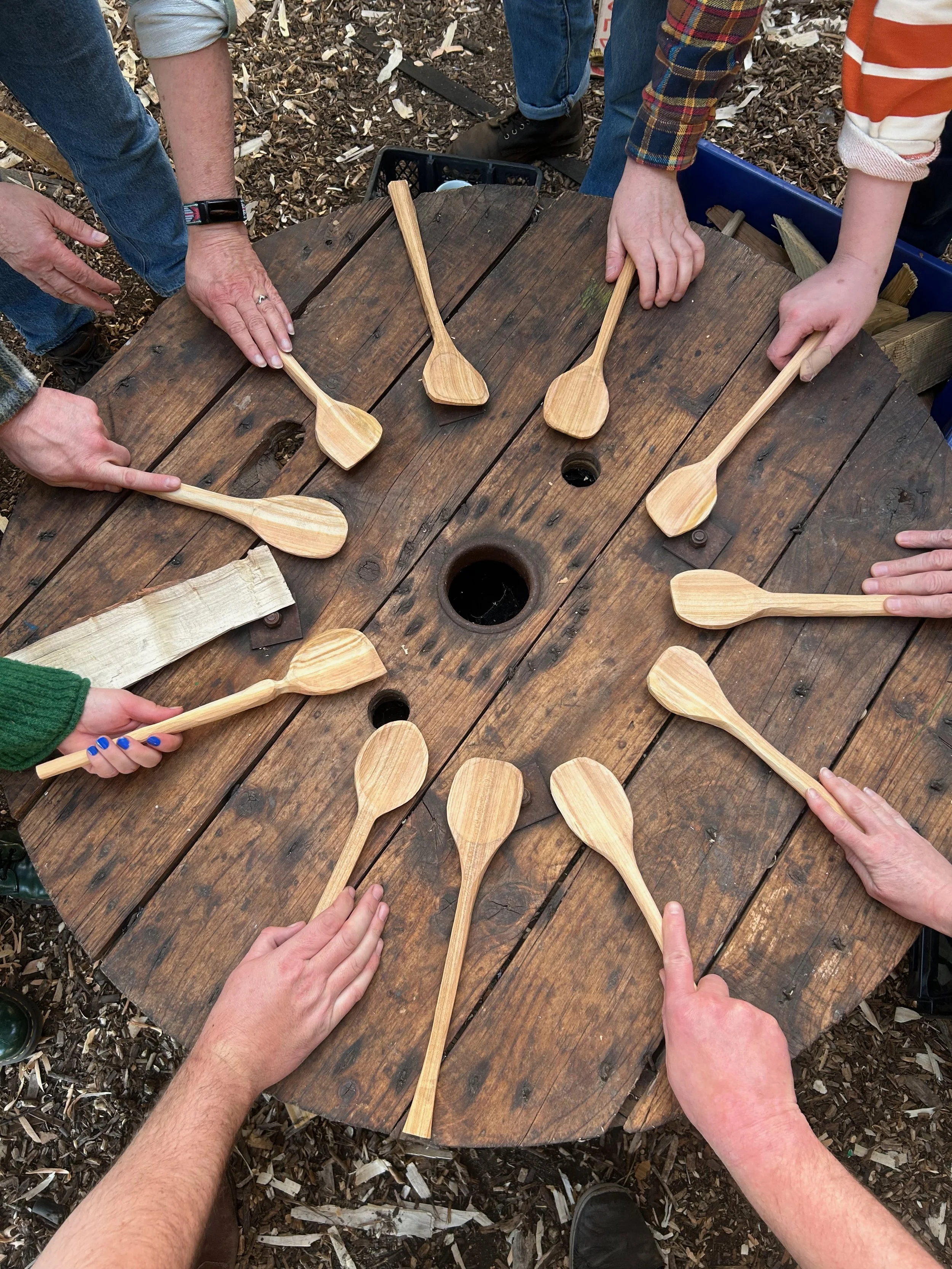 People gathered around a rustic wooden table holding unfinished wooden spoons.