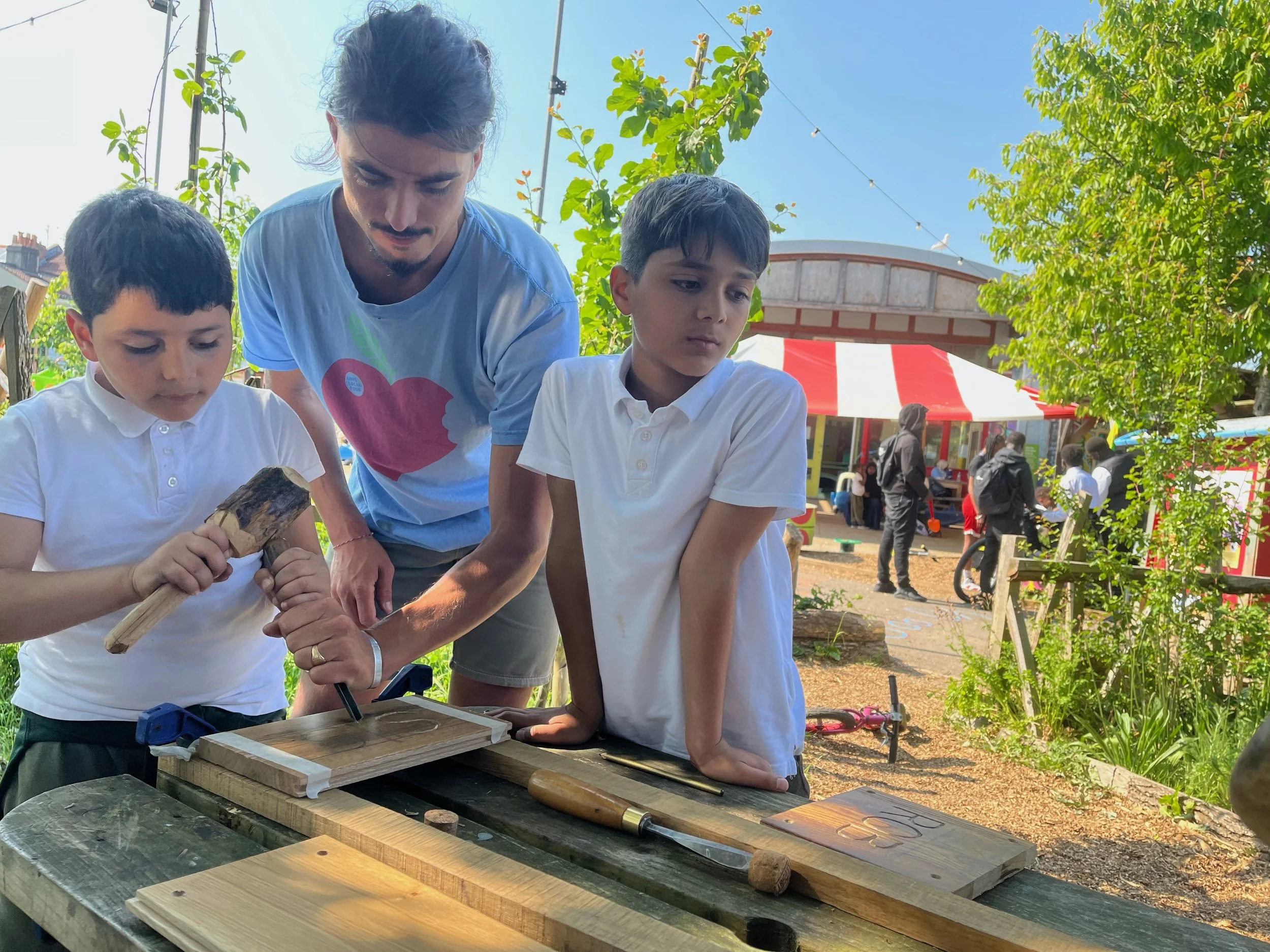 Two young boys working on a woodworking project outdoors, supervised by an adult man, at a craft fair or outdoor market with a red and white striped tent and people in the background.