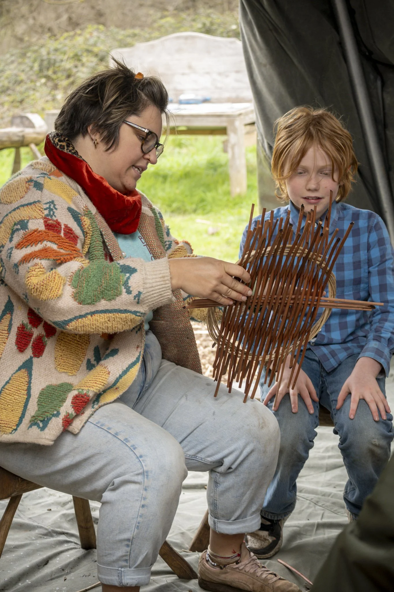 A woman with glasses and a colorful sweater is holding a small, rectangular, wooden frame with multiple thin sticks protruding from it, while a young boy with red hair and a plaid shirt watches attentively. They are outdoors, sitting on a wooden stoo