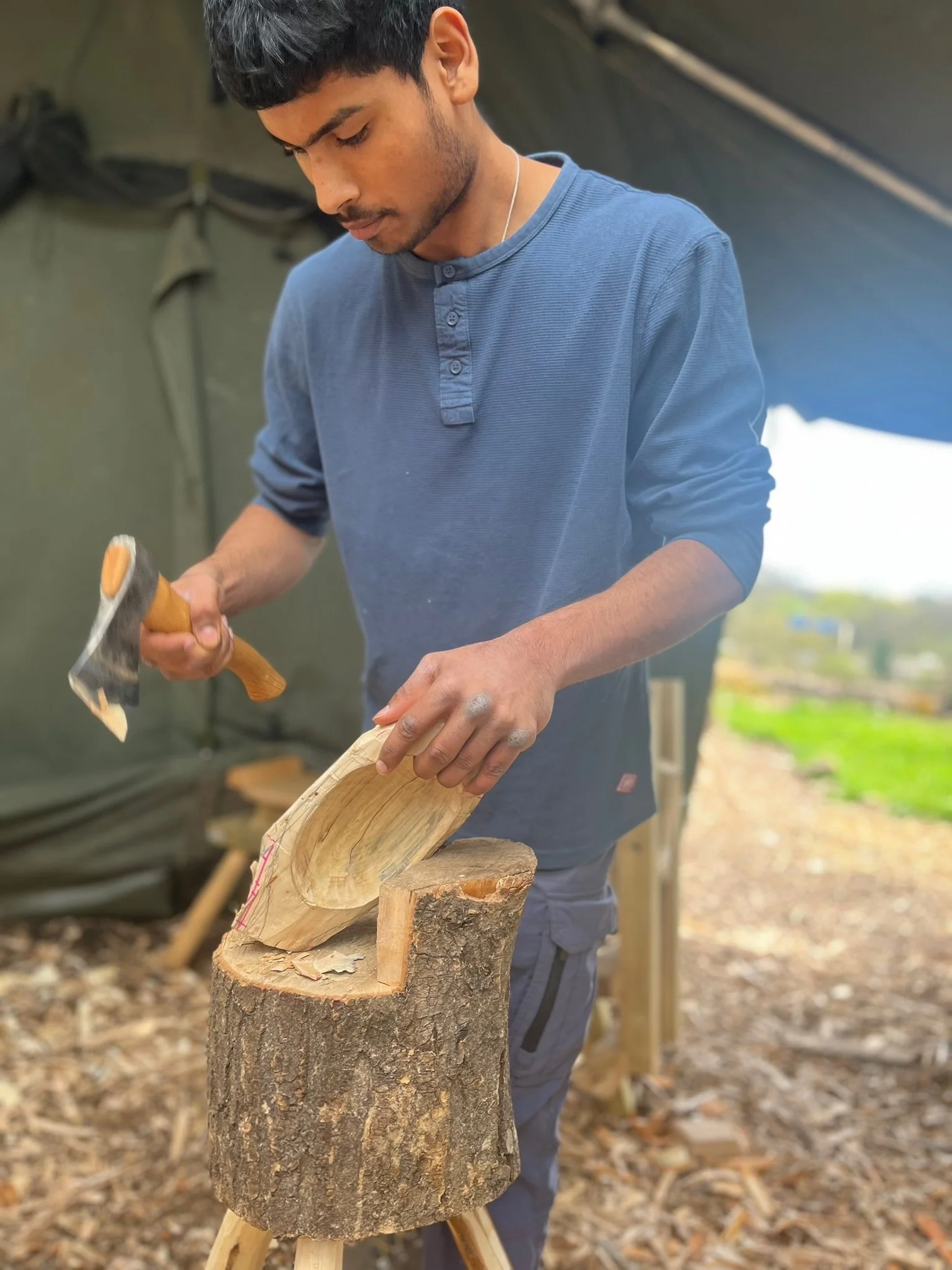 Young man carving a wooden sculpture with a hammer and chisel outdoors, standing over a tree stump used as a workbench.