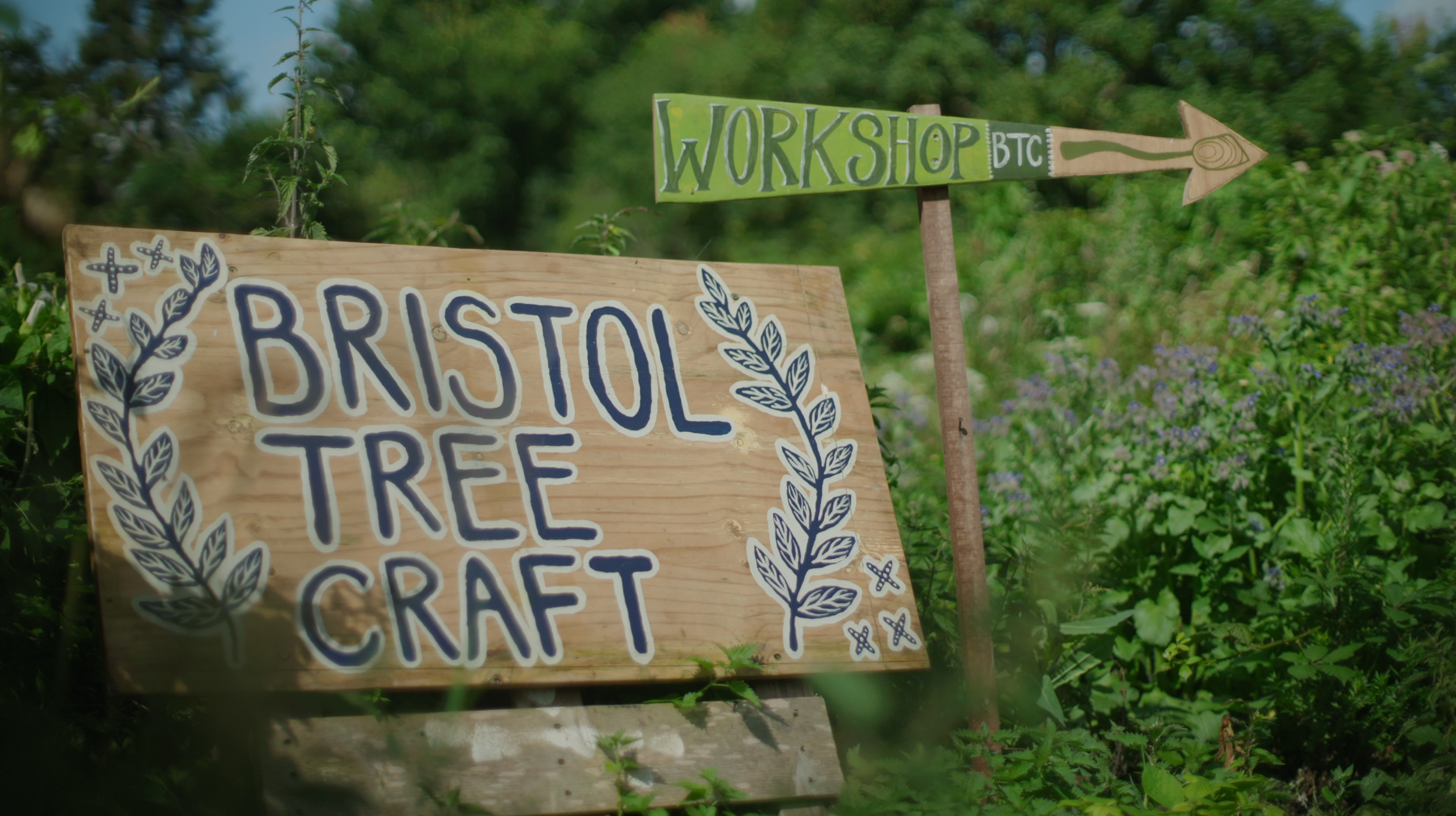 A wooden sign reads 'Bristol Tree Craft' decorated with blue leaf illustrations, situated among green foliage. A smaller sign with green and white lettering points to a workshop area, with an arrow indicating direction.